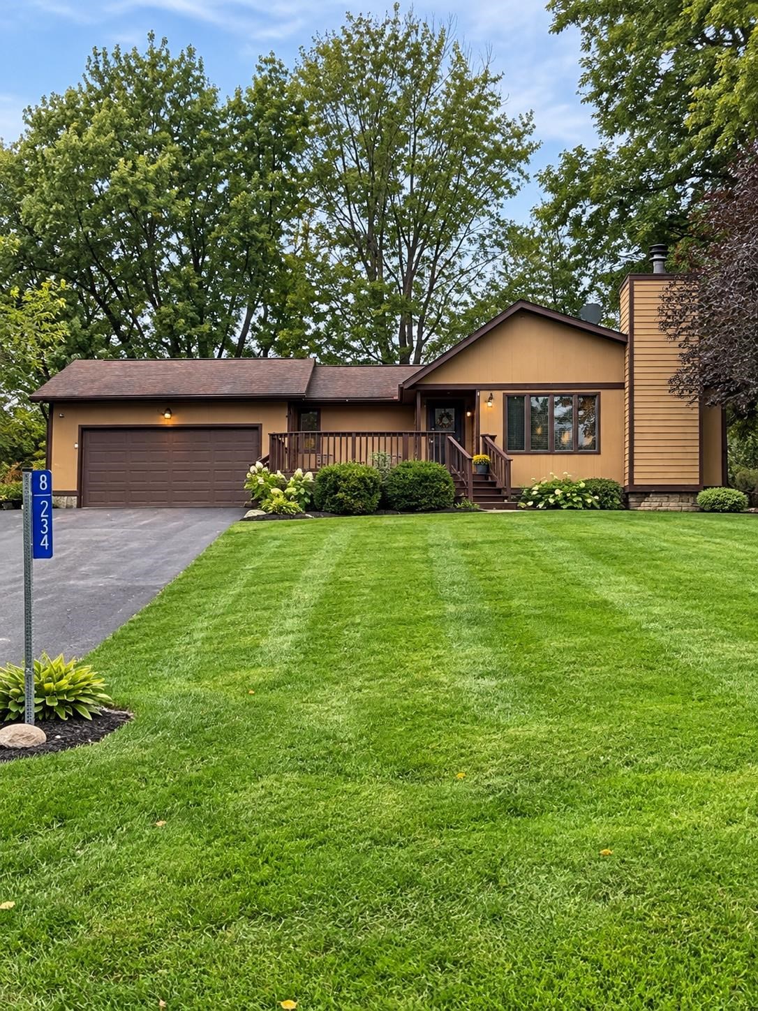 Tan single-story home with a double garage, raised front deck, and broad green lawn.