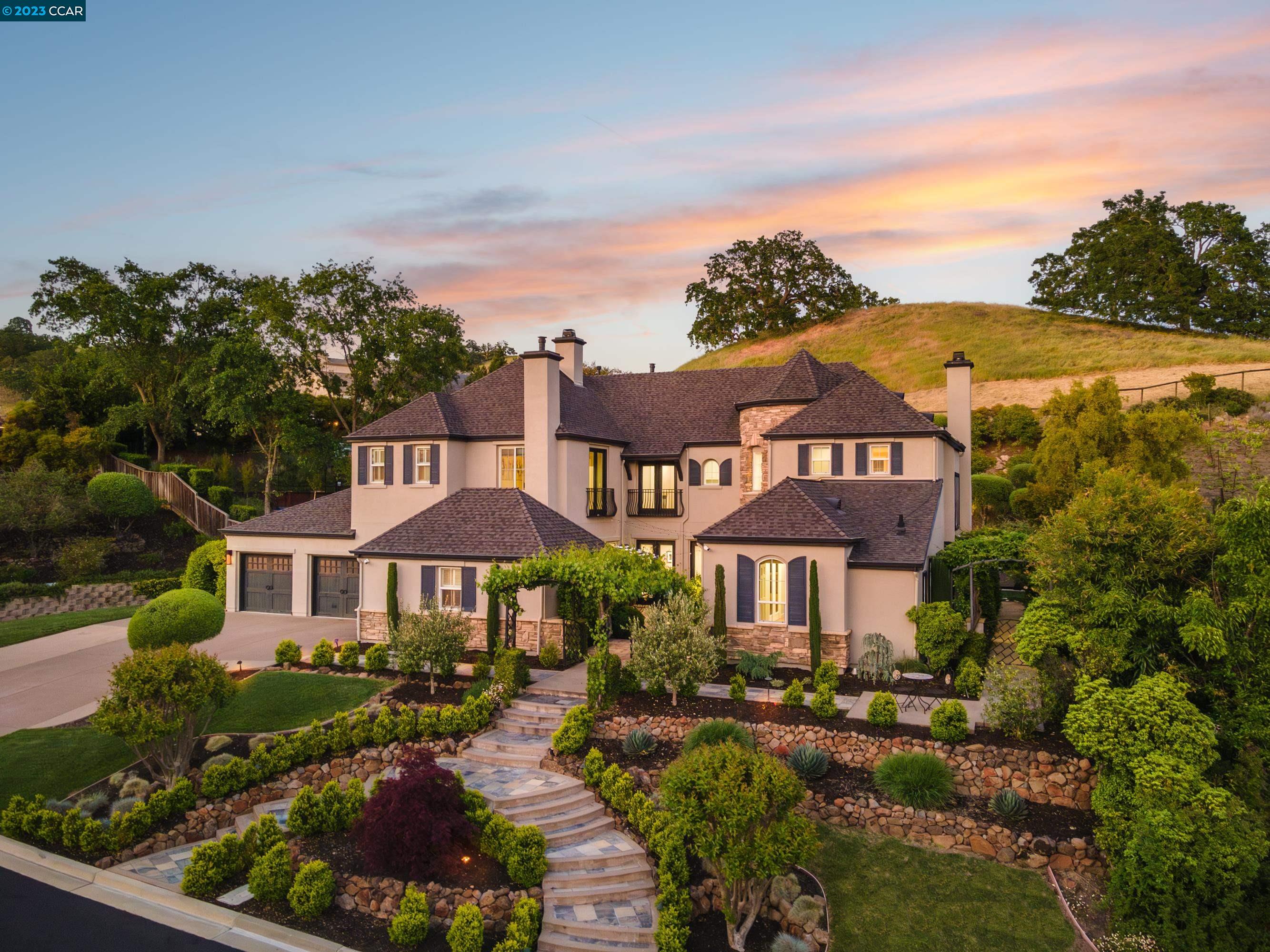 Two-story beige home with stone accents, landscaped gardens, and curved front steps at sunset.
