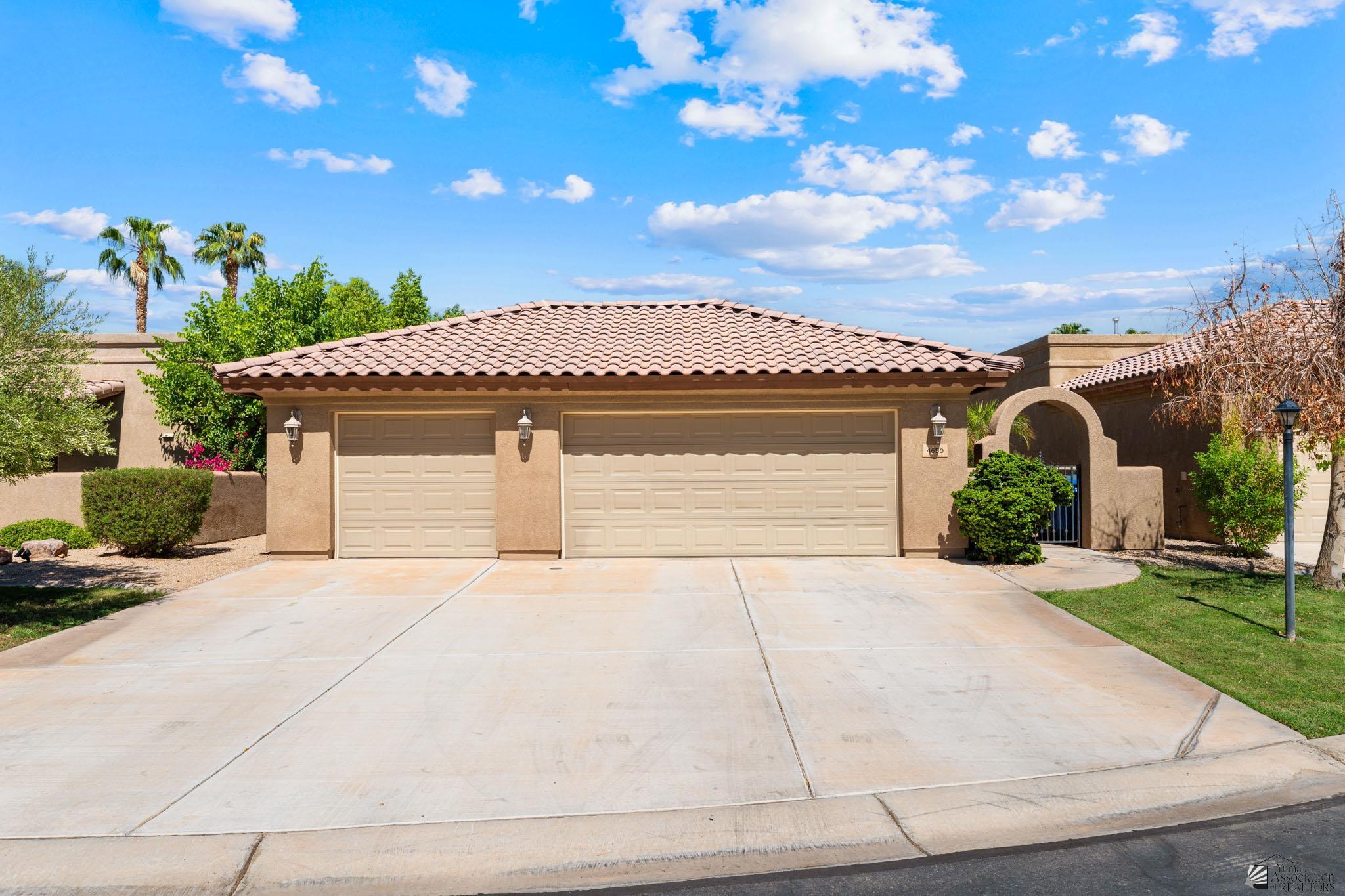 Tan stucco home with a tile roof, double garage, arched entry gate, and wide driveway.