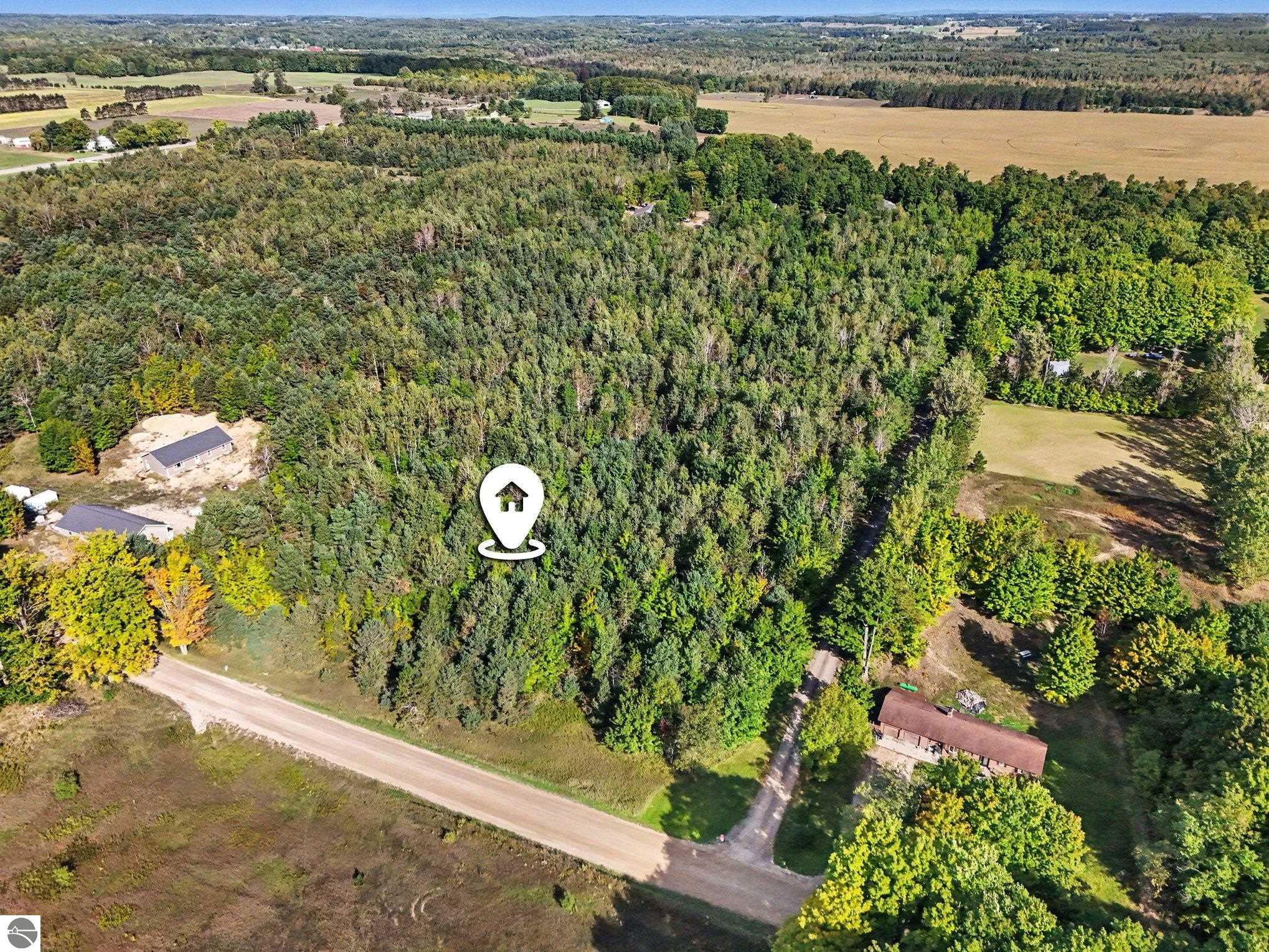Aerial view of wooded rural acreage with scattered buildings, open fields, and a road along its edge.