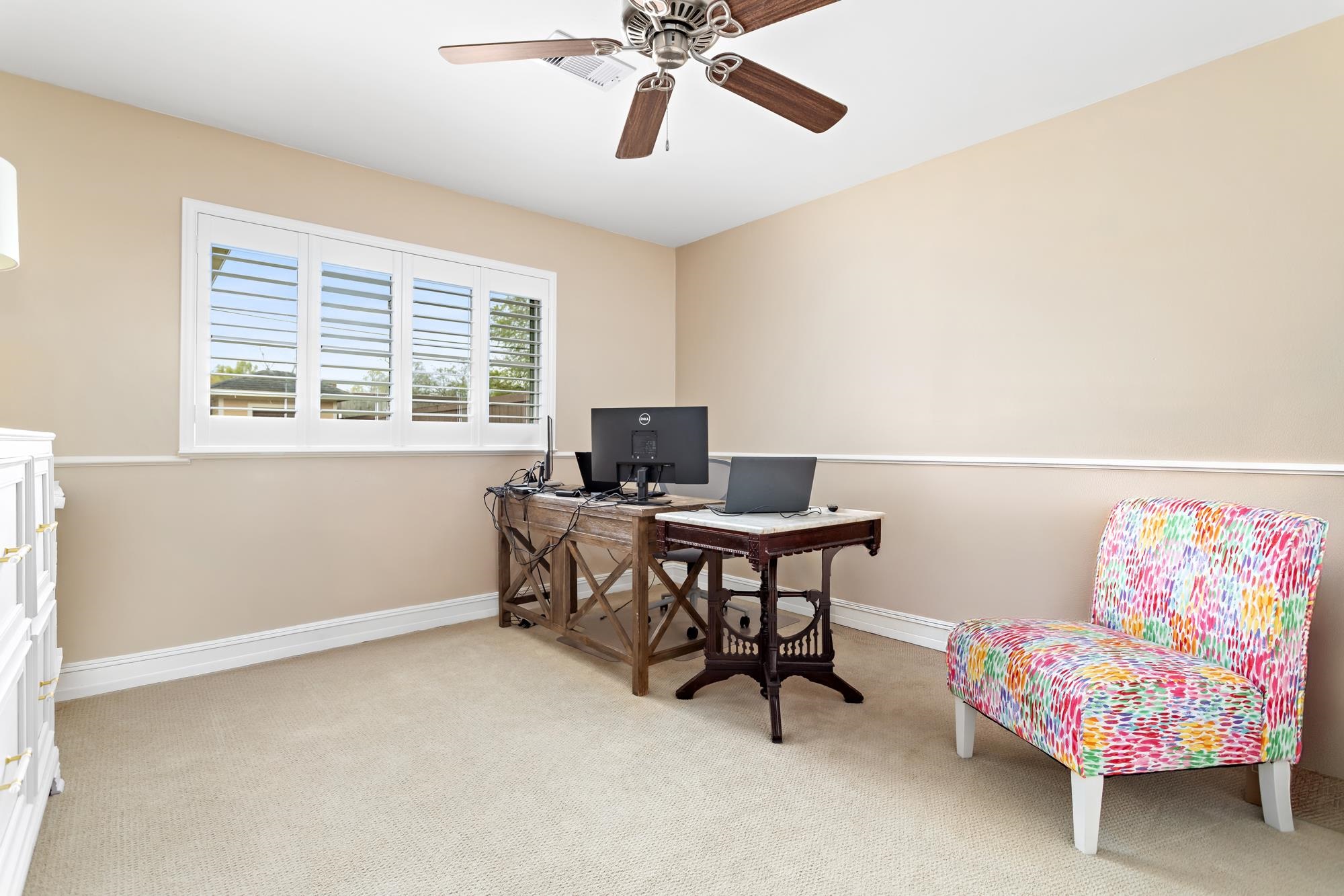 Home office with wooden desk, colorful accent chair, and plantation shutters