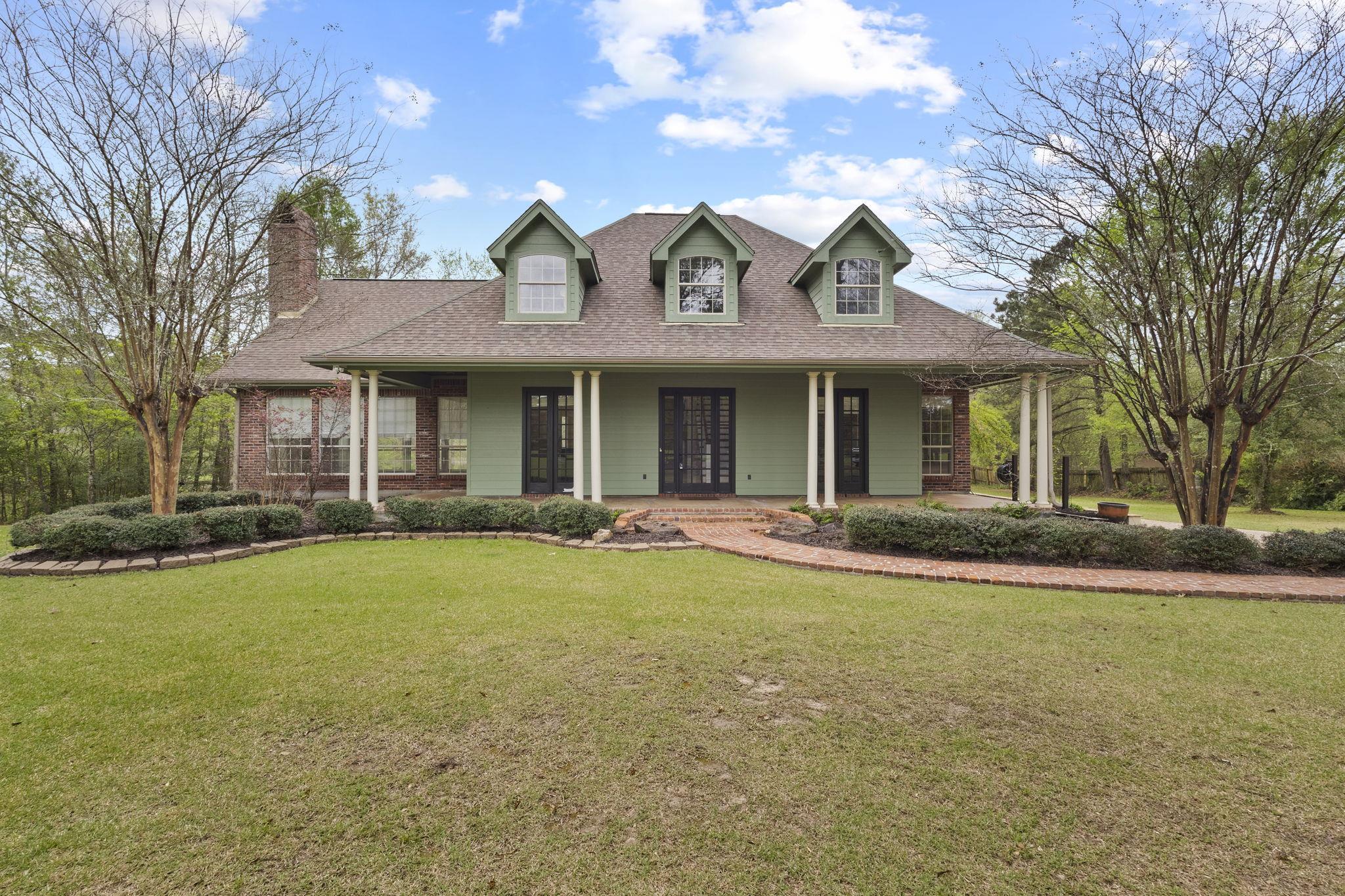Front view of single-story ranch home with green siding, brick accents, wraparound porch with white columns, and dormer windows