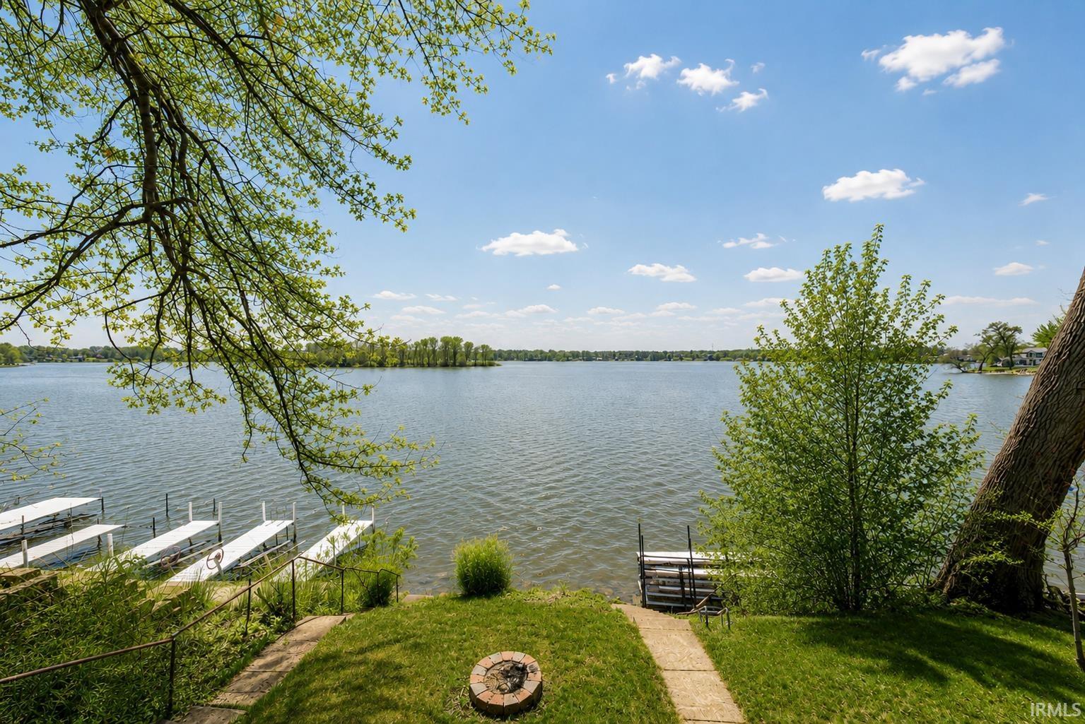 Expansive waterfront property featuring multiple private boat docks, a concrete path to the water's edge, a built-in stone fire pit, and mature trees