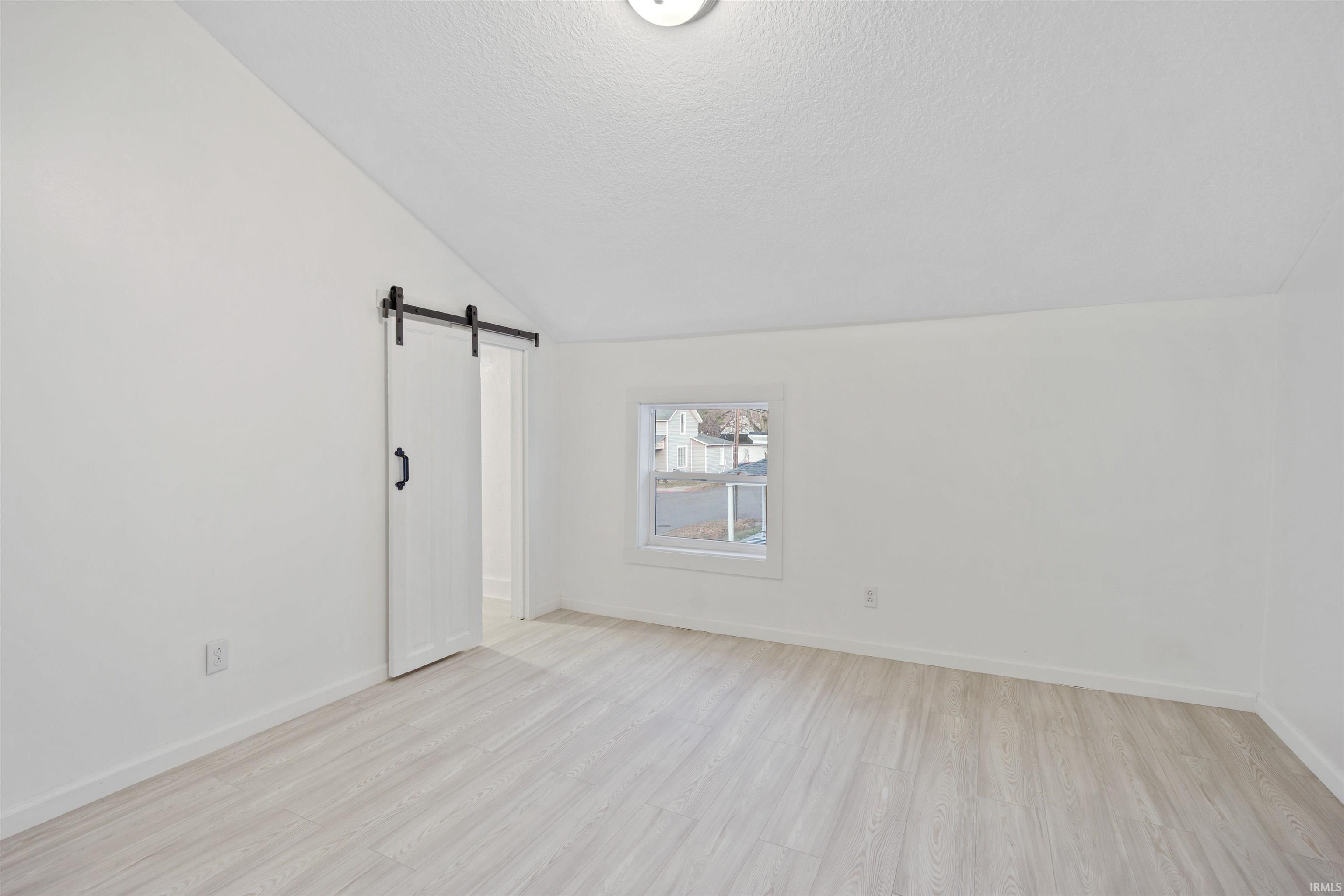 Empty room with a barn door, vaulted ceiling, light wood finished floors, and a textured ceiling