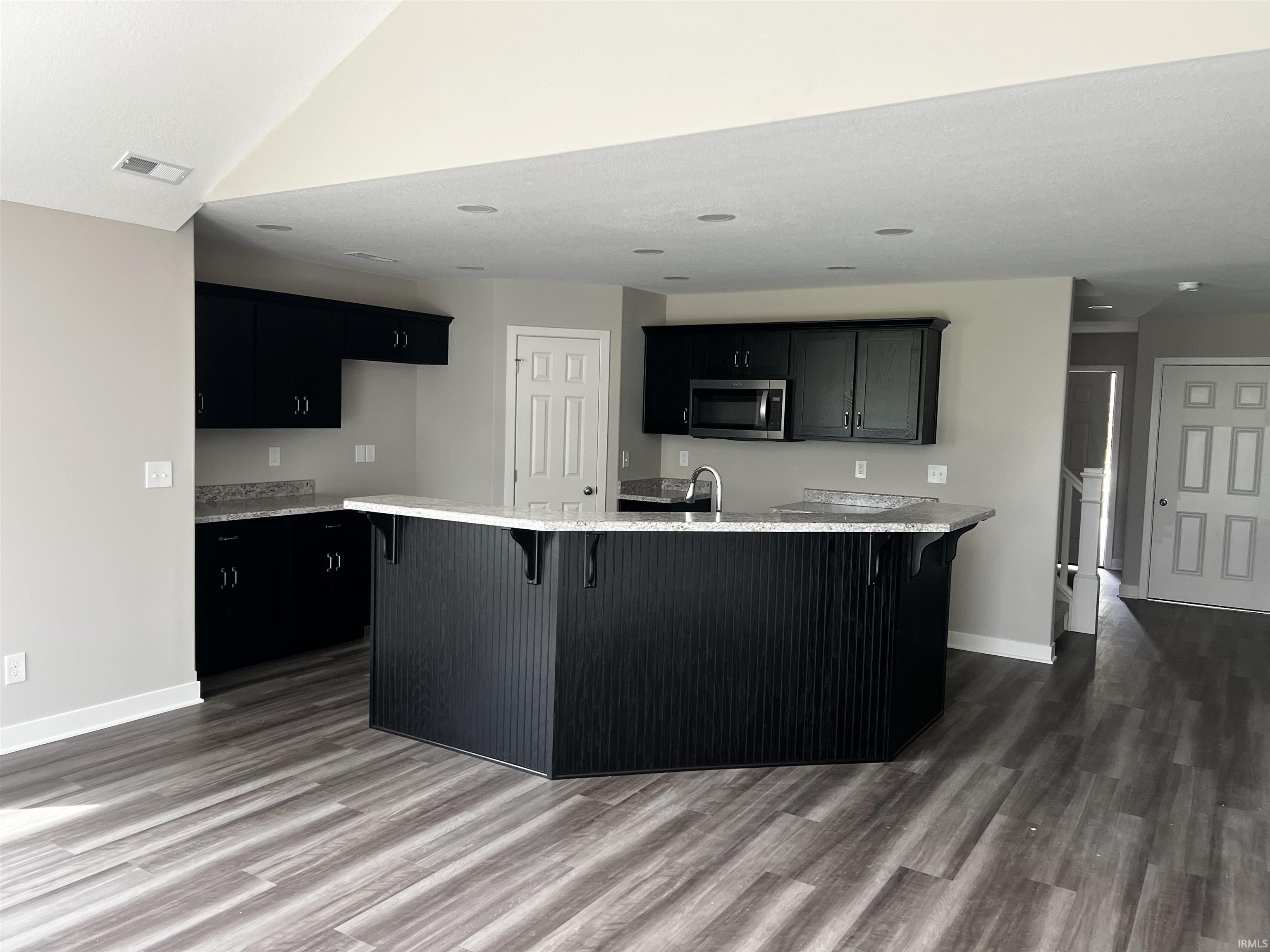 Kitchen with a kitchen bar, dark cabinets, light stone counters, stainless steel microwave, and dark wood-style flooring