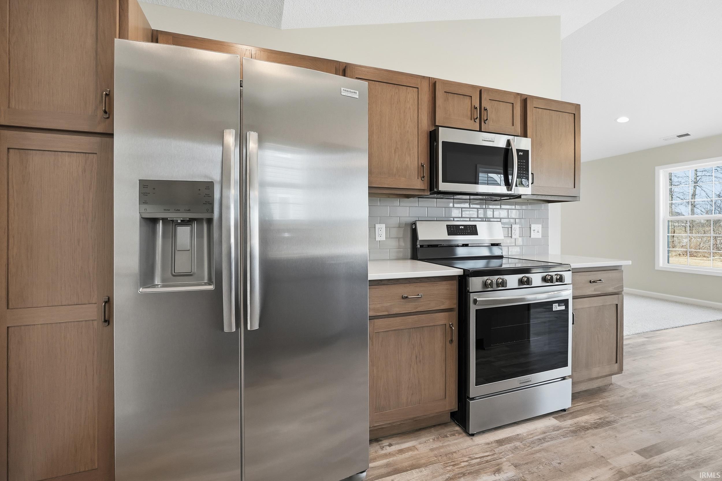Kitchen with stainless steel appliances, lofted ceiling, light wood finished floors, brown cabinetry, and decorative backsplash