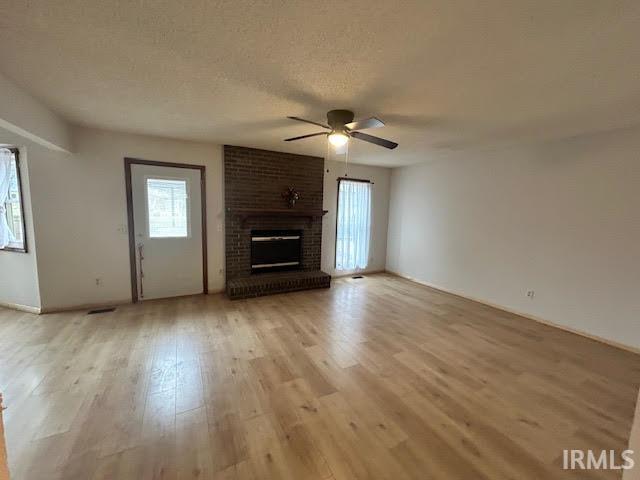 Unfurnished living room with a brick fireplace, a textured ceiling, ceiling fan, light wood-style floors, and plenty of natural light