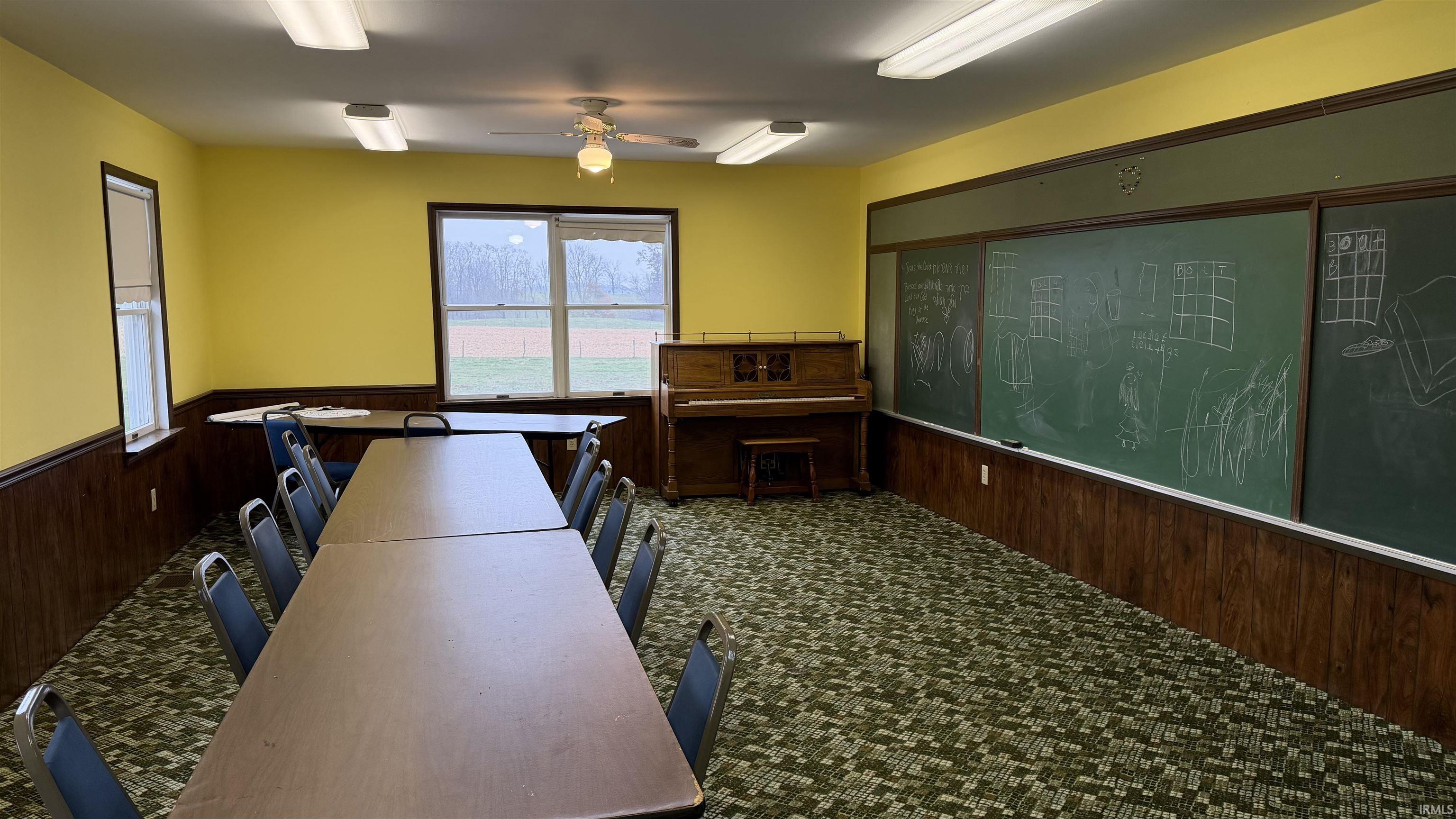 Carpeted classroom featuring wood walls, a wainscoted wall, huge chalk board and a ceiling fan.