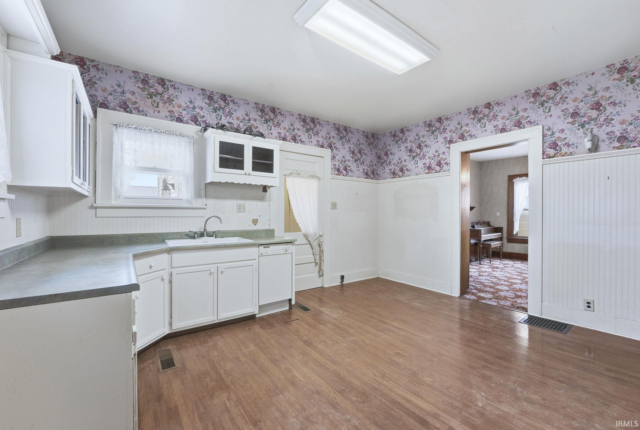 Kitchen with white cabinetry, wallpapered walls, dark wood-style flooring, glass insert cabinets, and white dishwasher