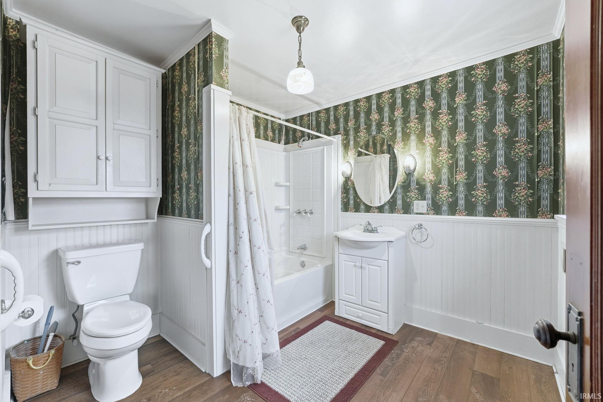 Full bath featuring vanity, shower / bath combo, dark wood finished floors, wallpapered walls, and wainscoting