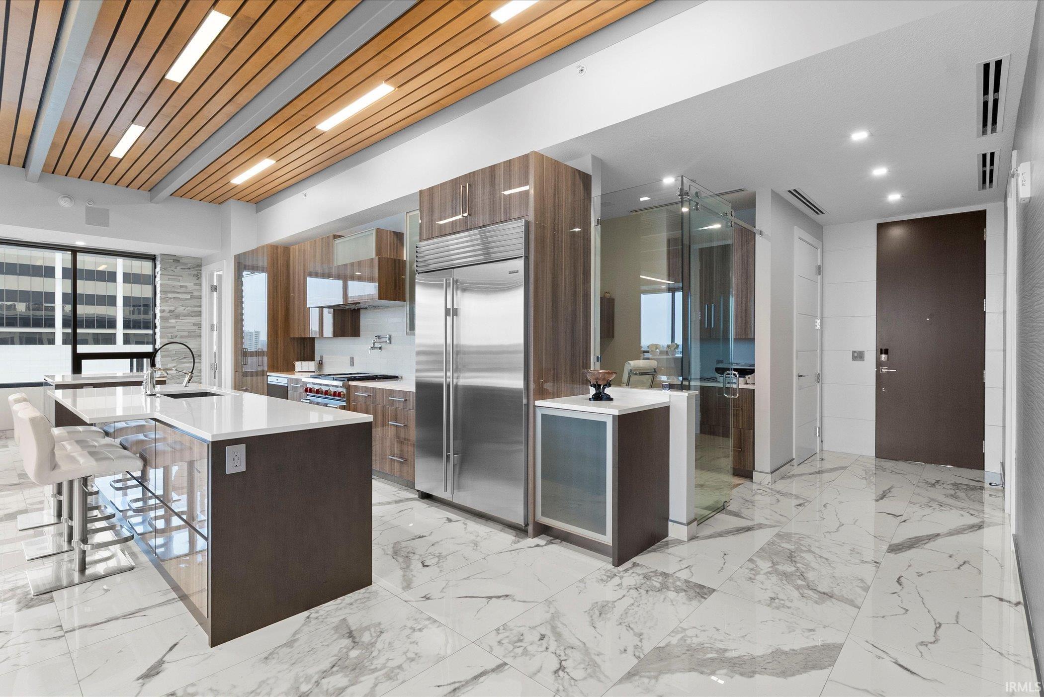 Kitchen featuring modern cabinets, light stone countertops, an island with sink, built in fridge, and a kitchen breakfast bar