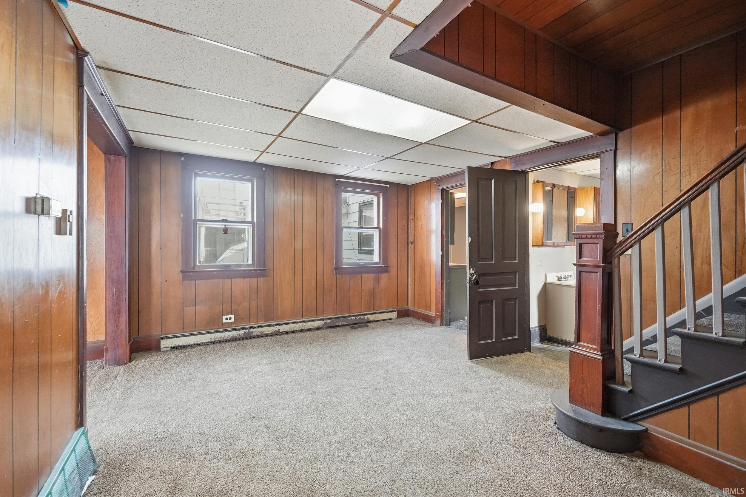 Below grade area with wood walls, light colored carpet, a drop ceiling, baseboard heating, and stairs