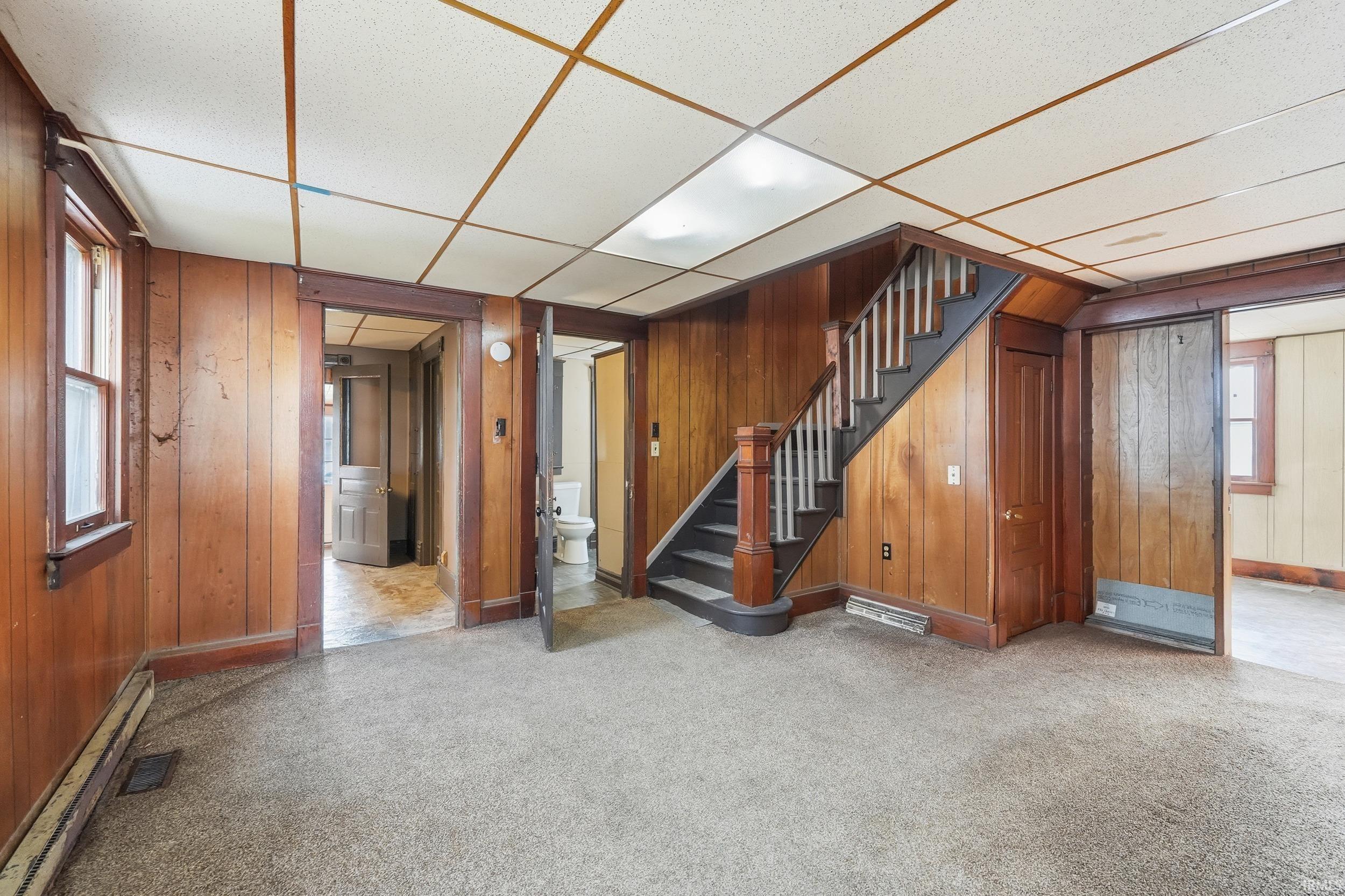 Basement featuring wooden walls, a drop ceiling, a baseboard heating unit, carpet floors, and stairs