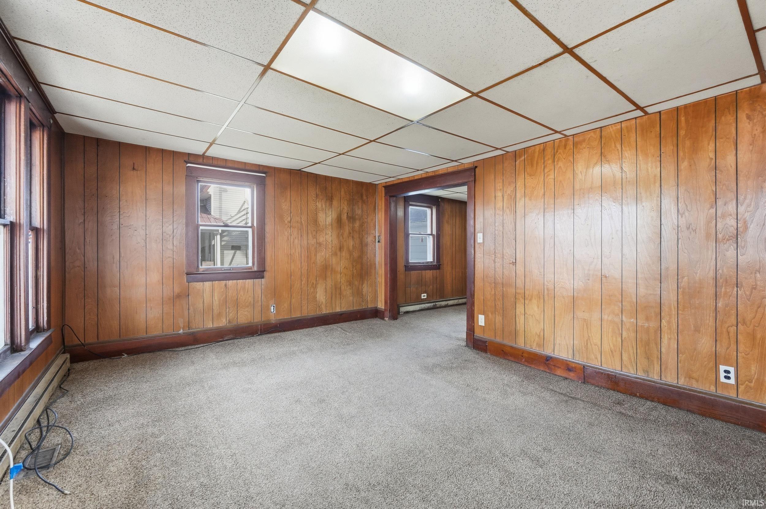 Carpeted empty room featuring a drop ceiling, a baseboard heating unit, and wood walls