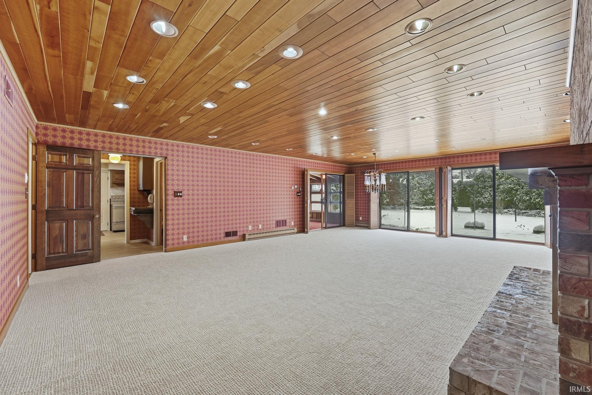 Unfurnished living room with wallpapered walls, wooden ceiling, carpet, a chandelier, and a baseboard heating unit