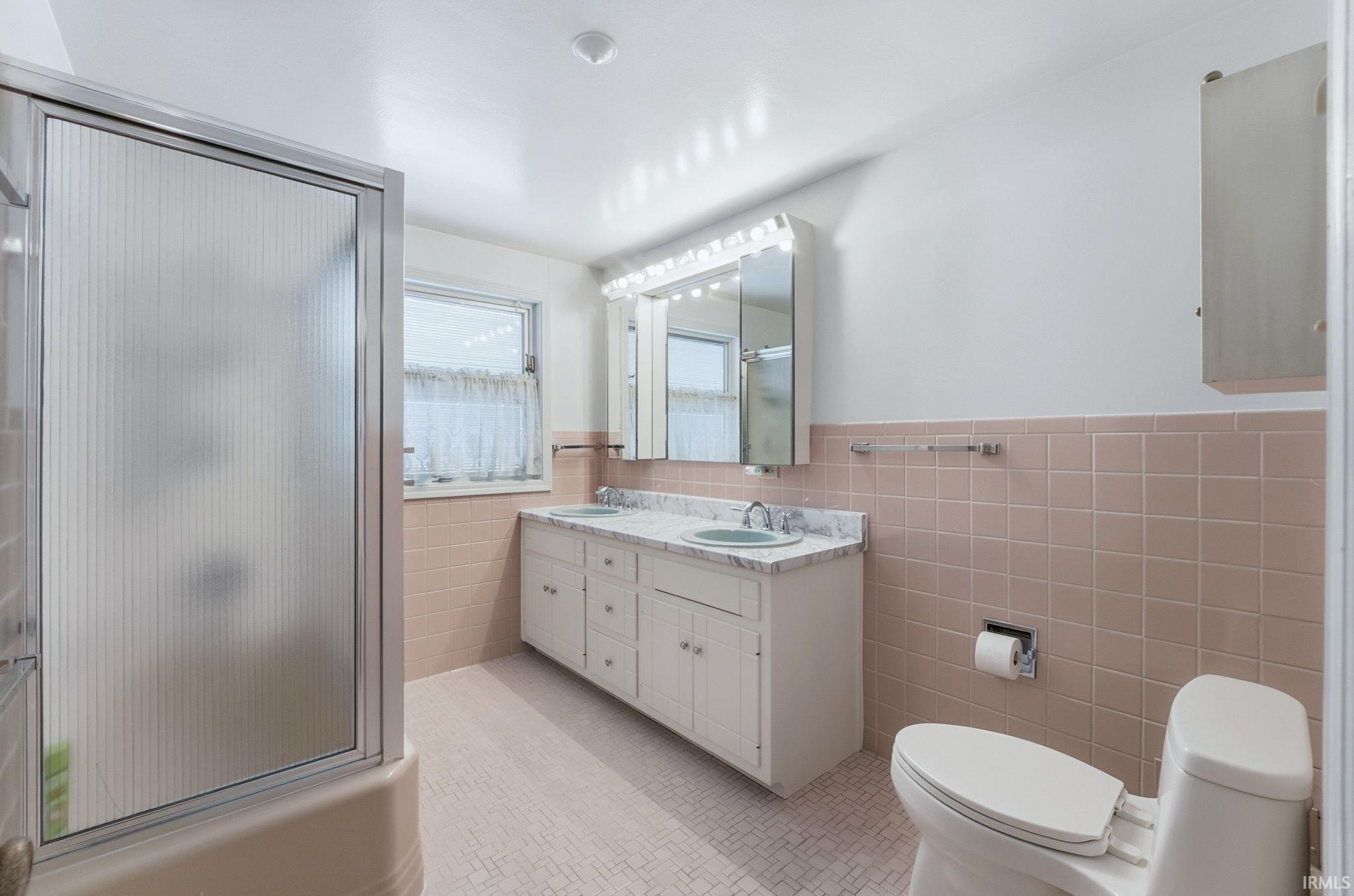 Bathroom featuring a shower stall, double vanity, wainscoting, tile walls, and light tile patterned floors