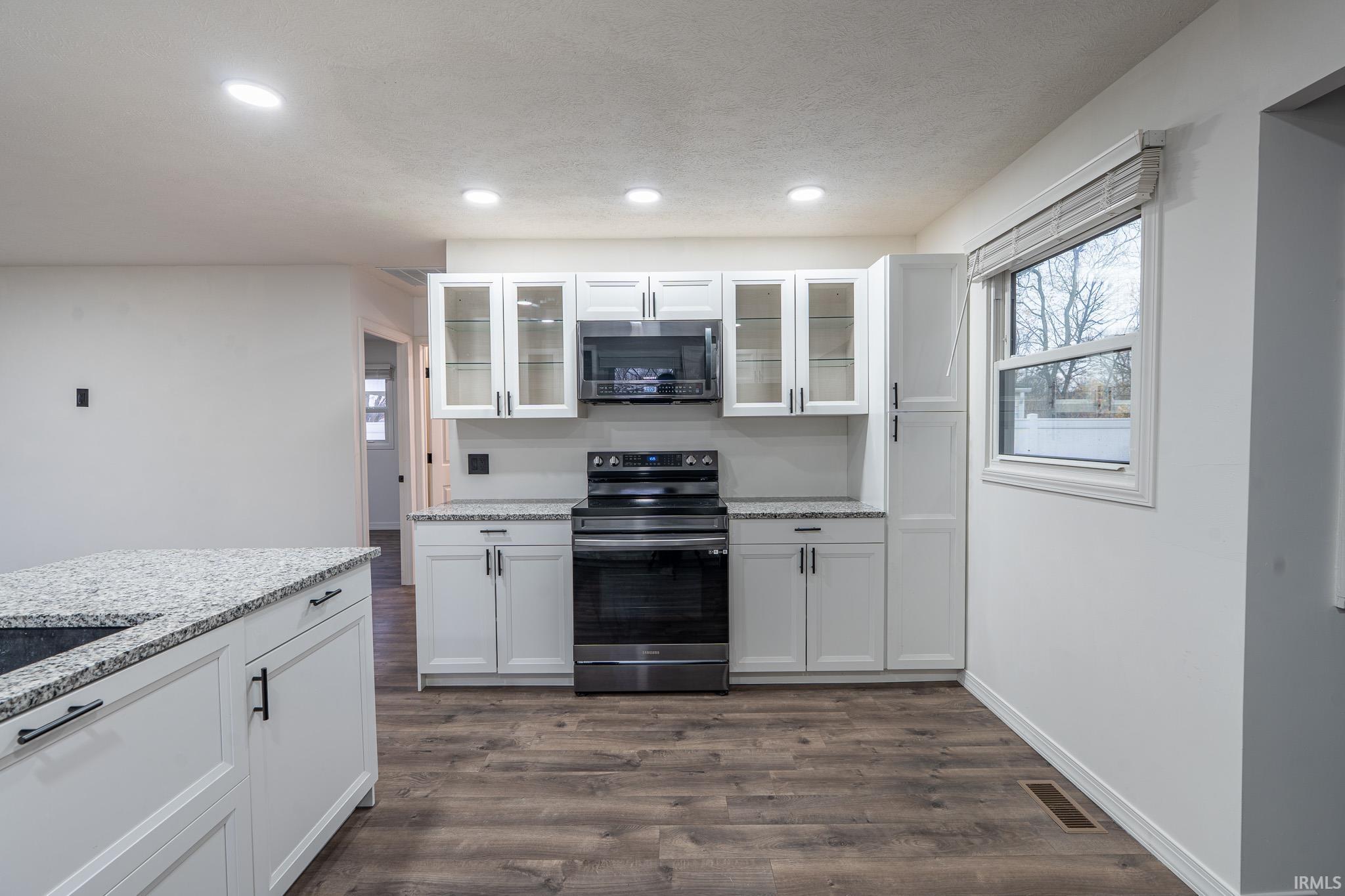 Kitchen with white cabinetry, glass insert cabinets, stainless steel appliances, light stone countertops, and dark wood-style floors