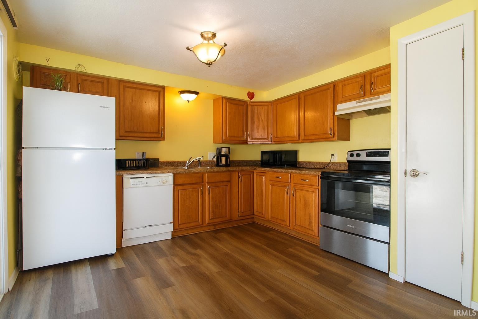 Kitchen with white appliances, brown cabinets, under cabinet range hood, dark countertops, and dark wood-style flooring