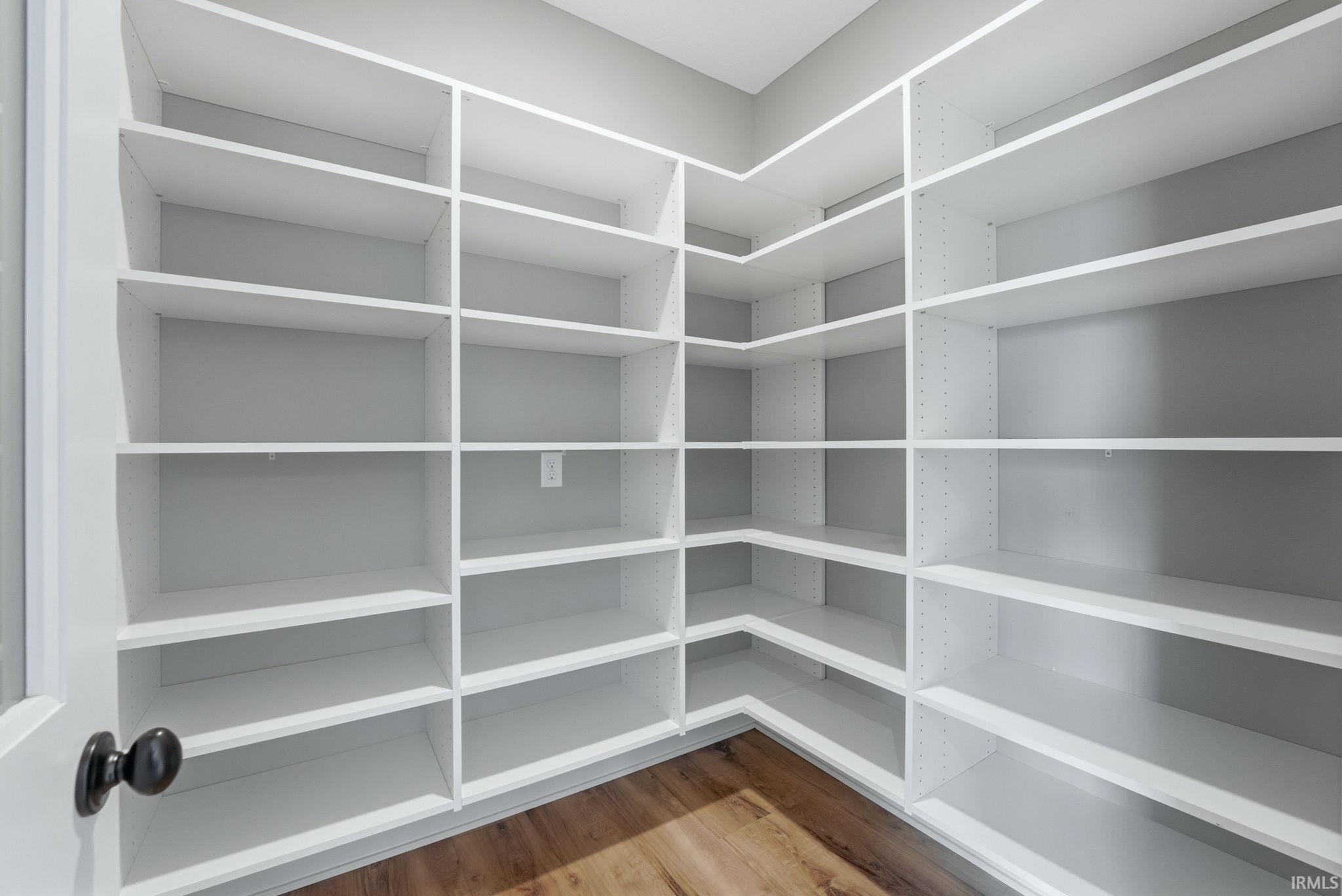 Expansive walk-in pantry featuring white built-in shelving, wood-finish flooring, and a contemporary black doorknob