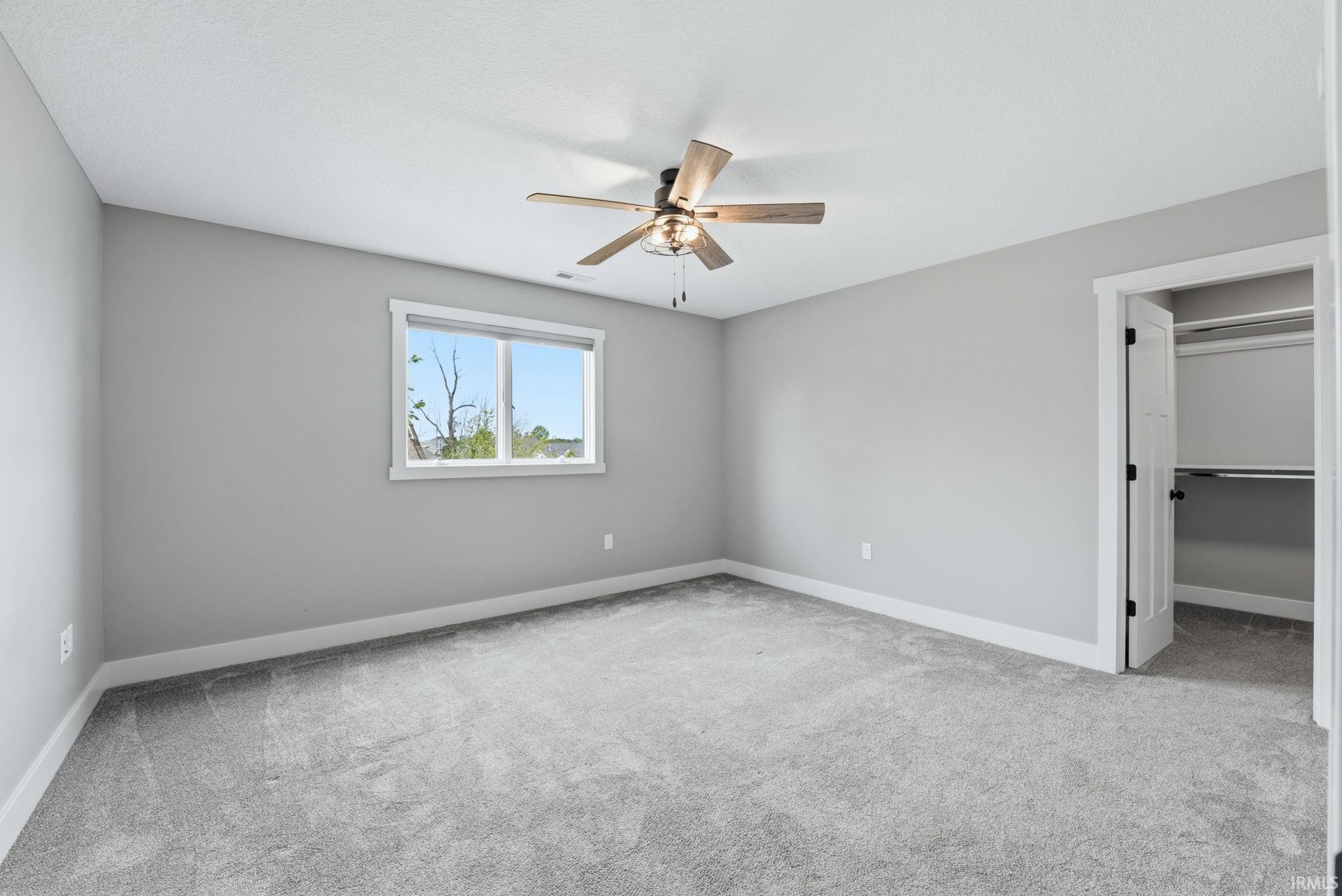 Spacious room featuring light gray walls, plush gray carpeting, a multi-pane window with white trim, a wood-finish ceiling fan, and a built-in closet with shelving and hanging rods