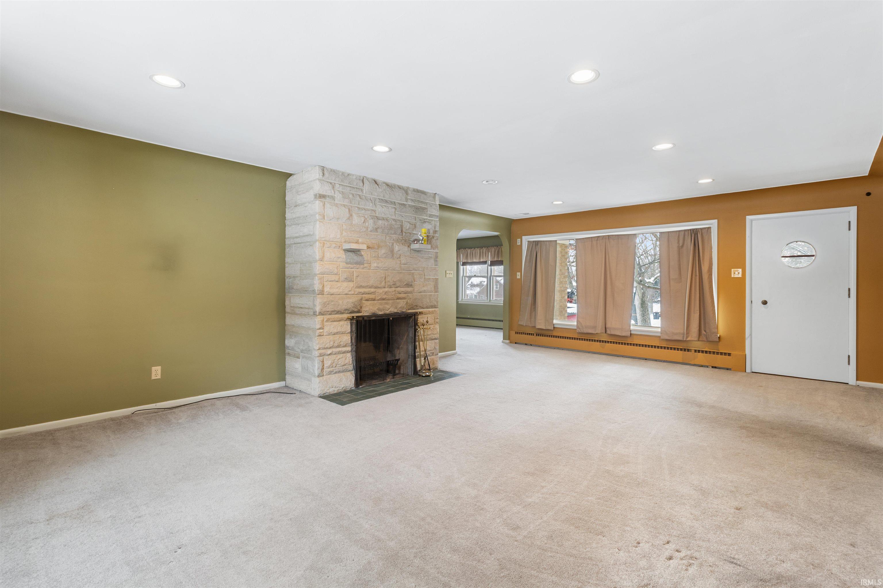 Unfurnished living room featuring arched walkways, a stone fireplace, a baseboard heating unit, carpet flooring, and recessed lighting