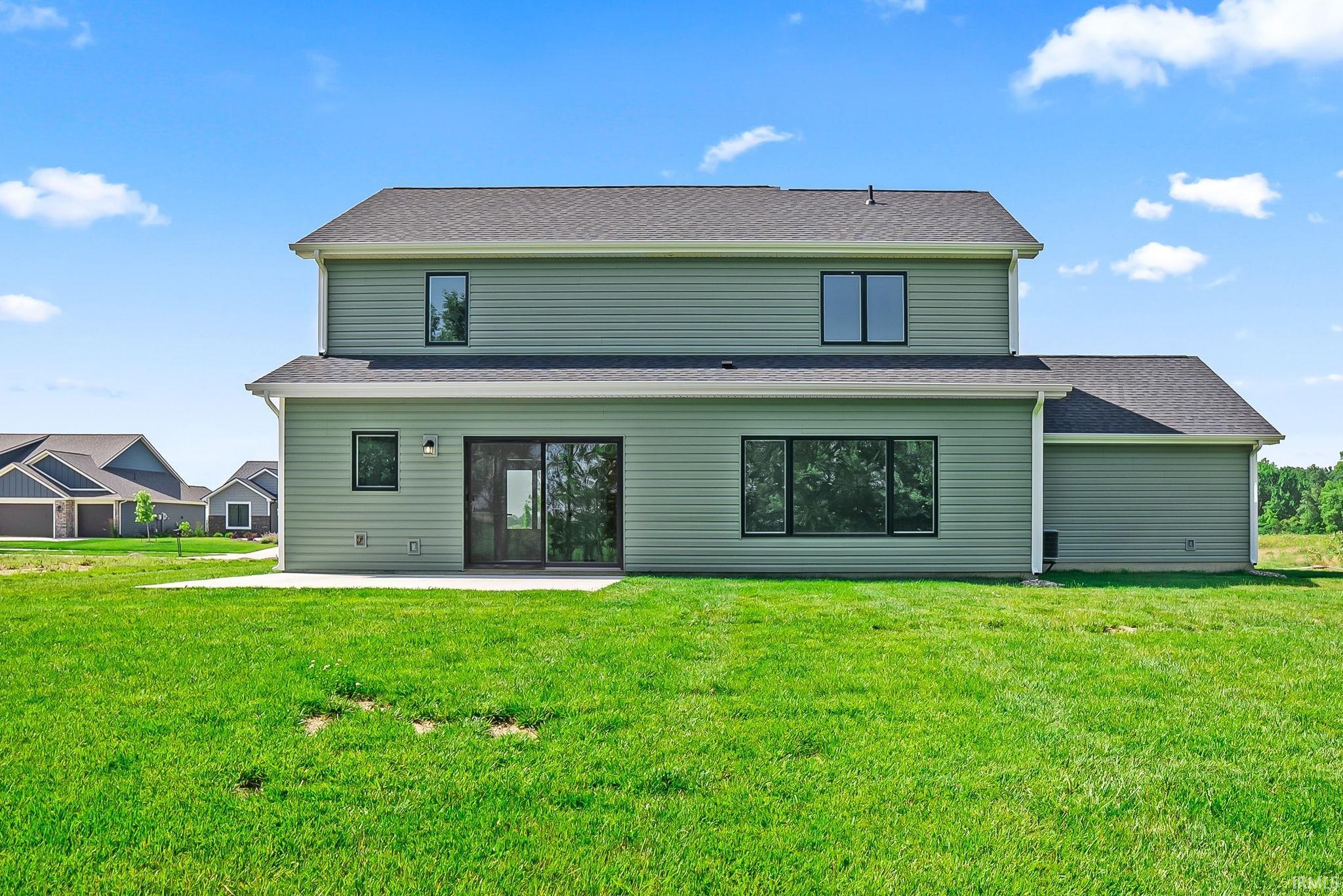 Spacious backyard featuring a concrete patio, light green horizontal siding, dark framed windows, a sliding glass door, and a dark shingled roof