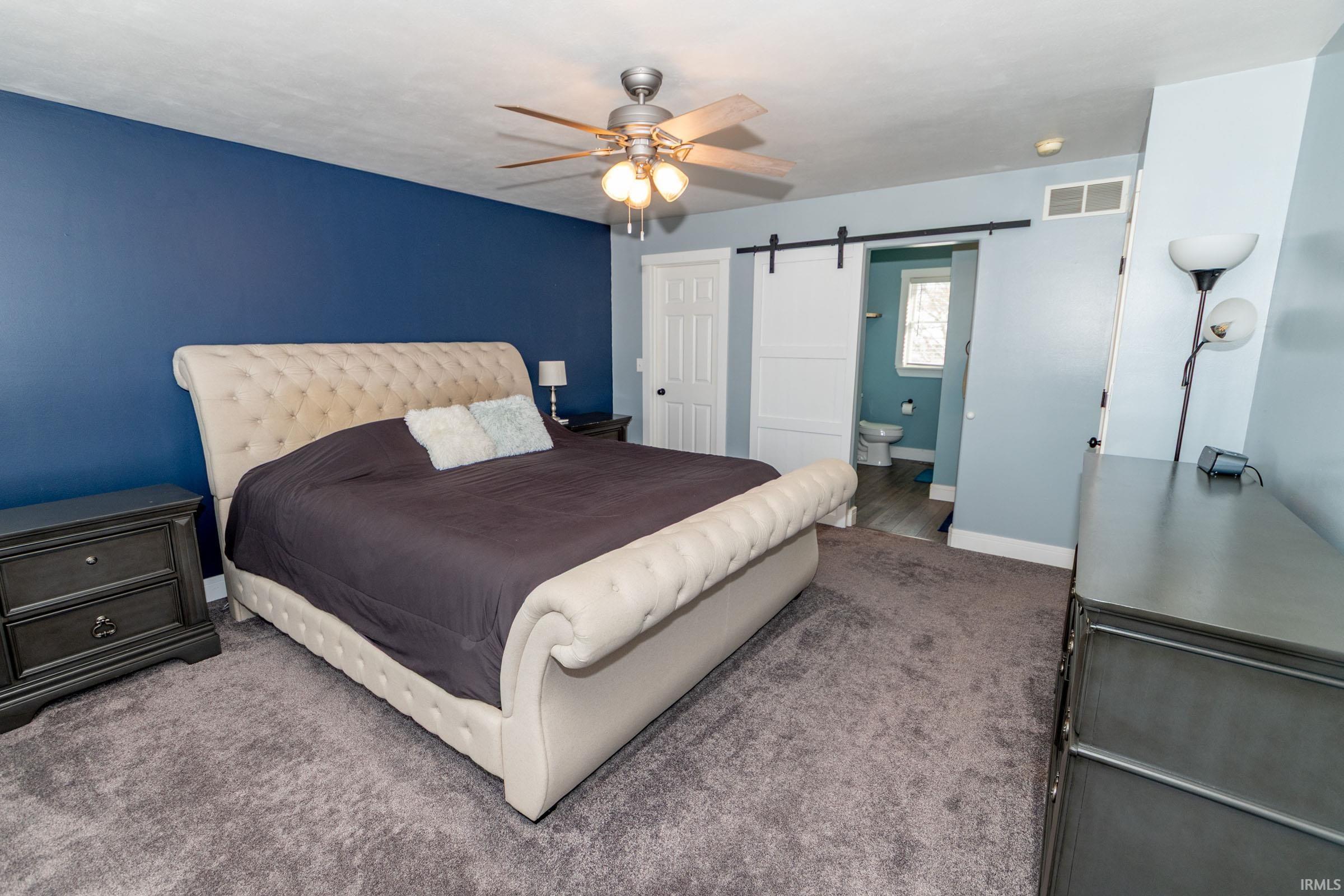 Bedroom featuring a barn door, a ceiling fan, ensuite bath, and carpet flooring