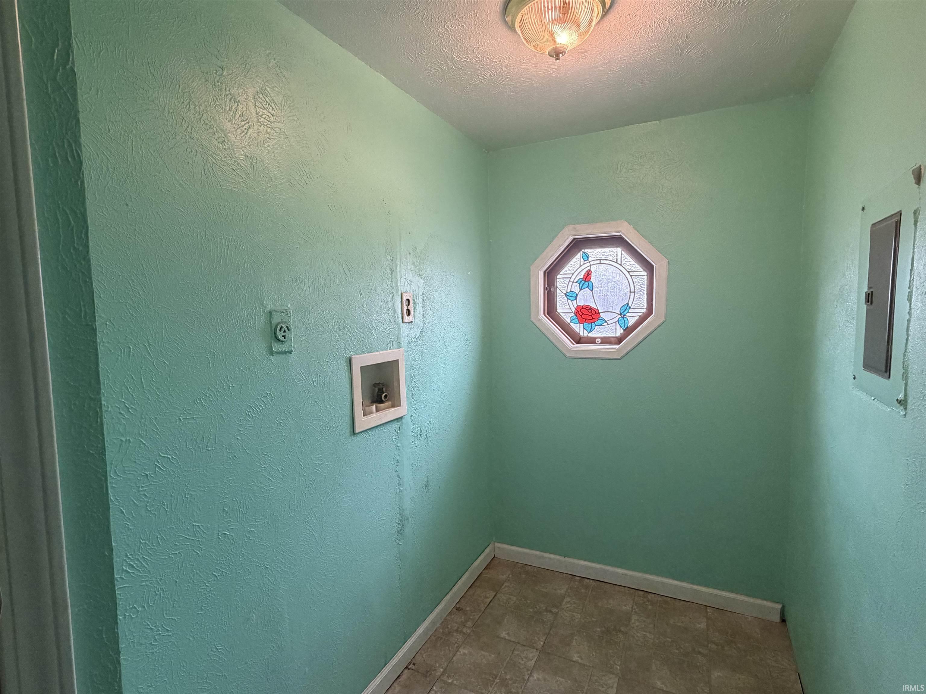 Laundry area featuring hookup for a washing machine, a textured ceiling, a textured wall, stone finish flooring, and electric panel