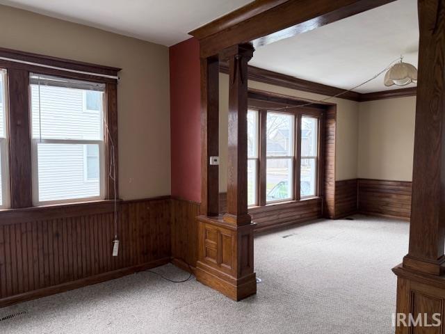 Empty room featuring wood walls, wainscoting, light colored carpet, and ornate columns