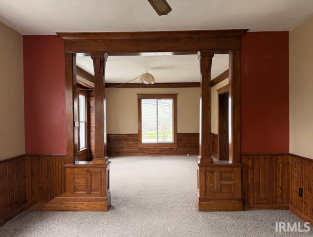 Empty room with wood walls, ornate columns, a wainscoted wall, carpet floors, and ceiling fan