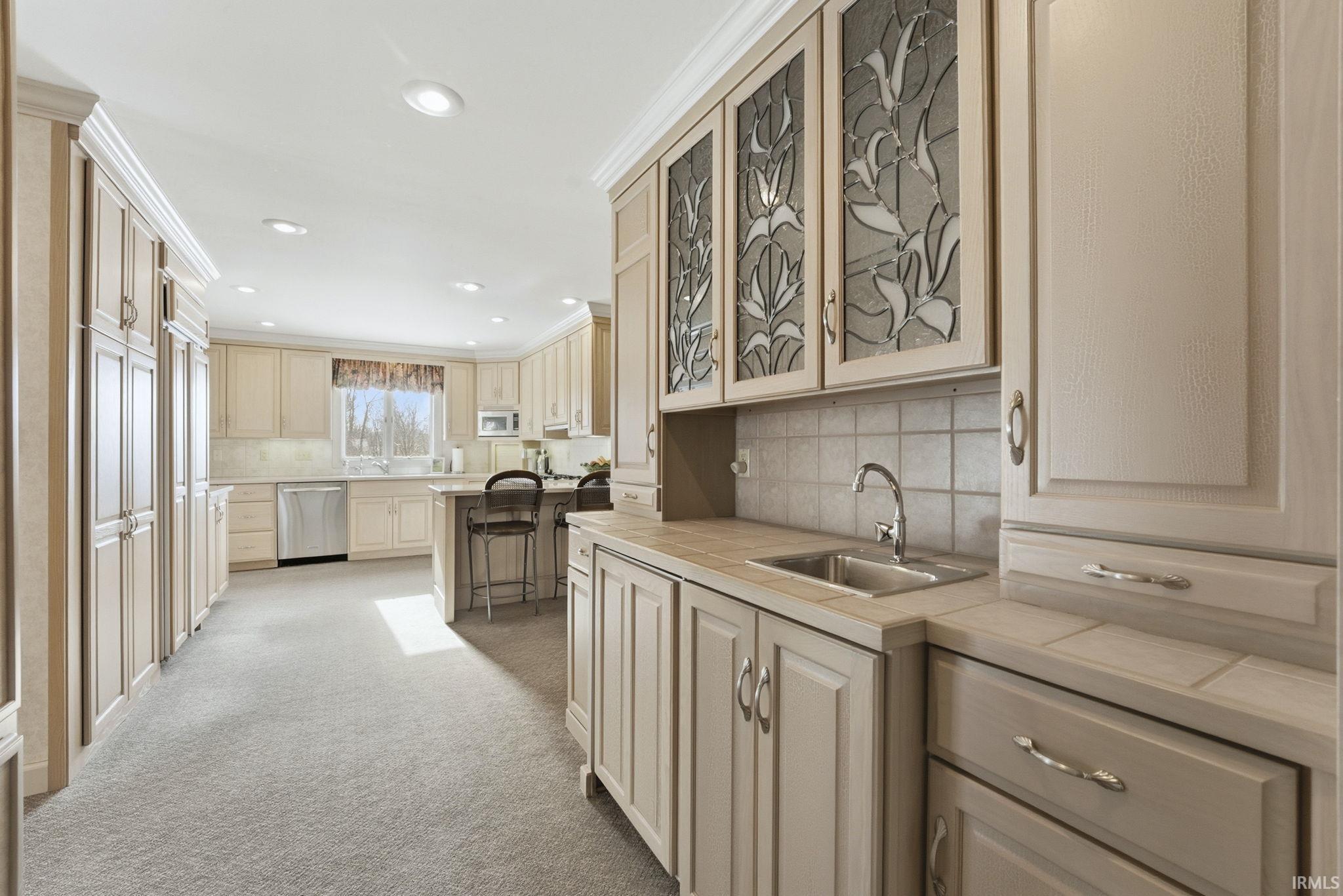 Kitchen featuring tile counters, glass insert cabinets, light carpet, ornamental molding, and decorative backsplash