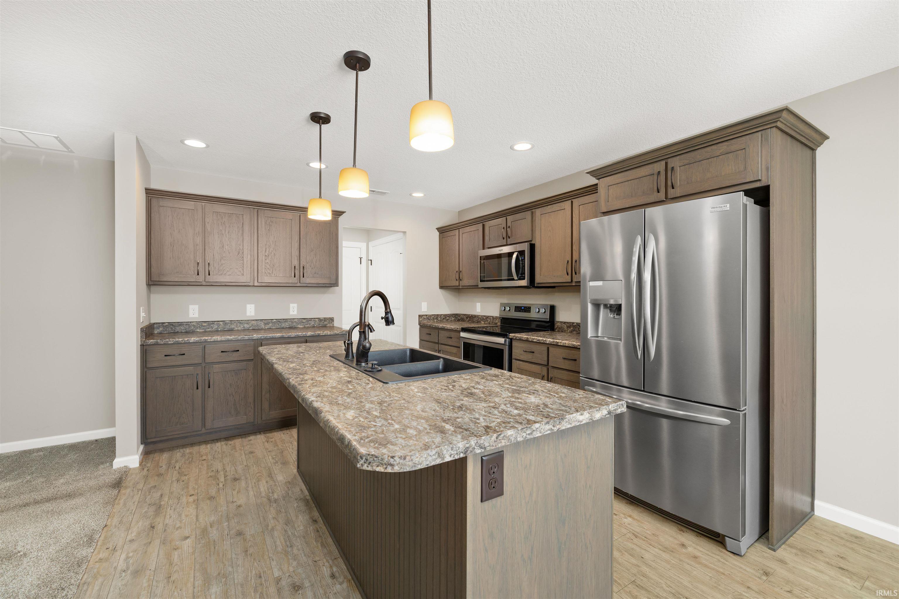 Kitchen featuring appliances with stainless steel finishes, an island with sink, hanging light fixtures, light countertops, and light wood-style floors