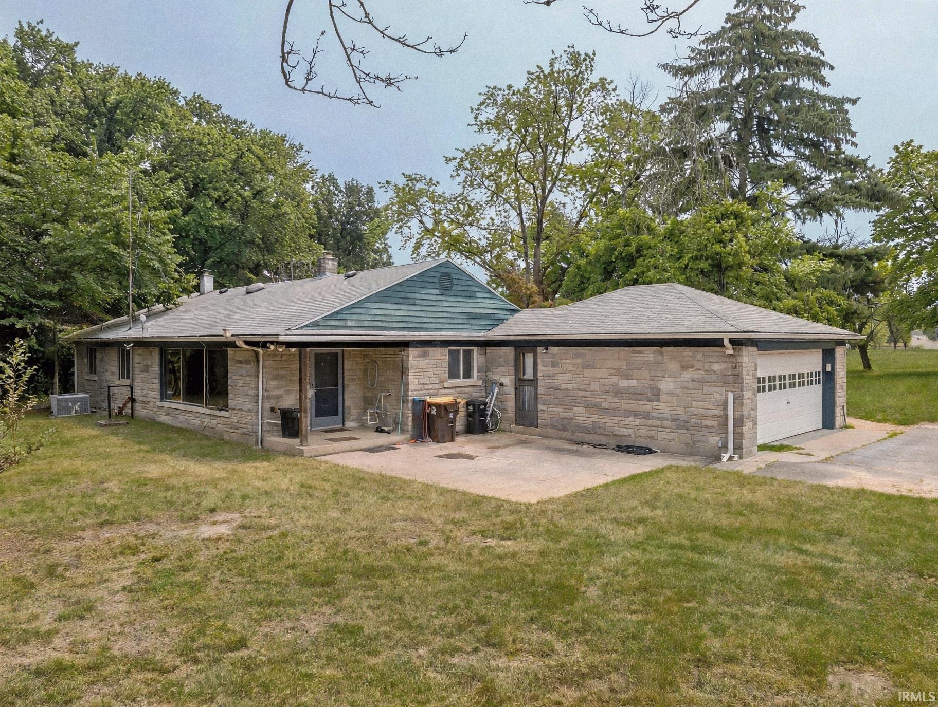Stone exterior residence featuring a covered patio, attached garage with a white sectional door, and mature surrounding trees