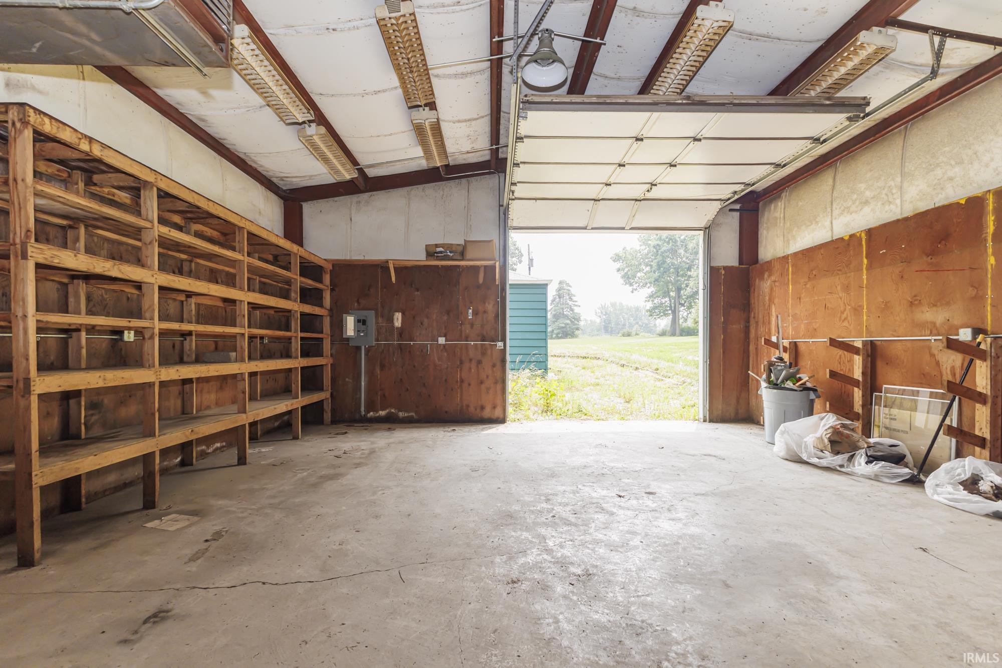 Spacious utility building featuring concrete flooring, extensive wooden shelving, an overhead door, interior lighting, and a visible electrical panel
