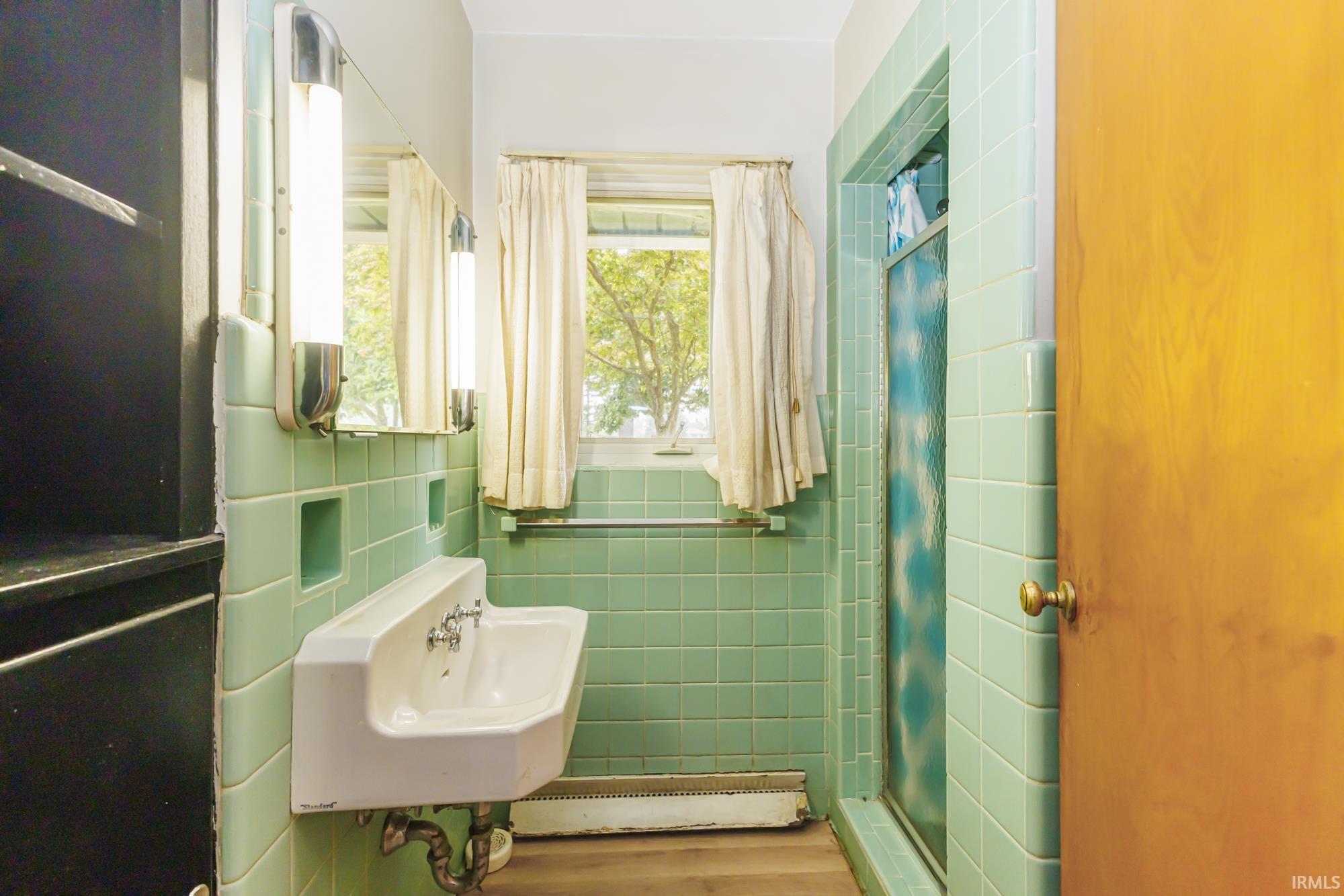 Bathroom featuring light green square tile walls, a white wall-mounted sink with dual faucets, and a shower with a patterned glass door