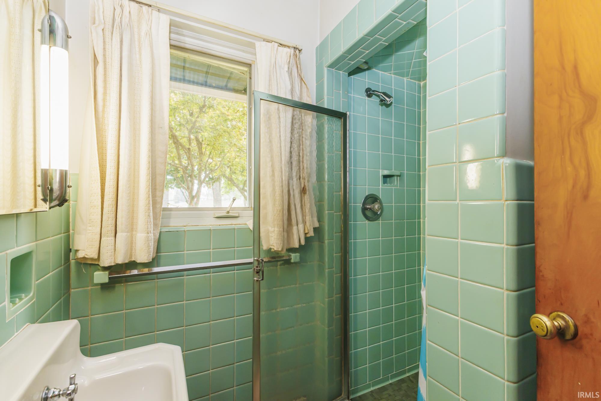 Bathroom featuring mint green tiled walls, a built-in shower with a glass enclosure, a window, a wall-mounted sink, and a chrome towel bar