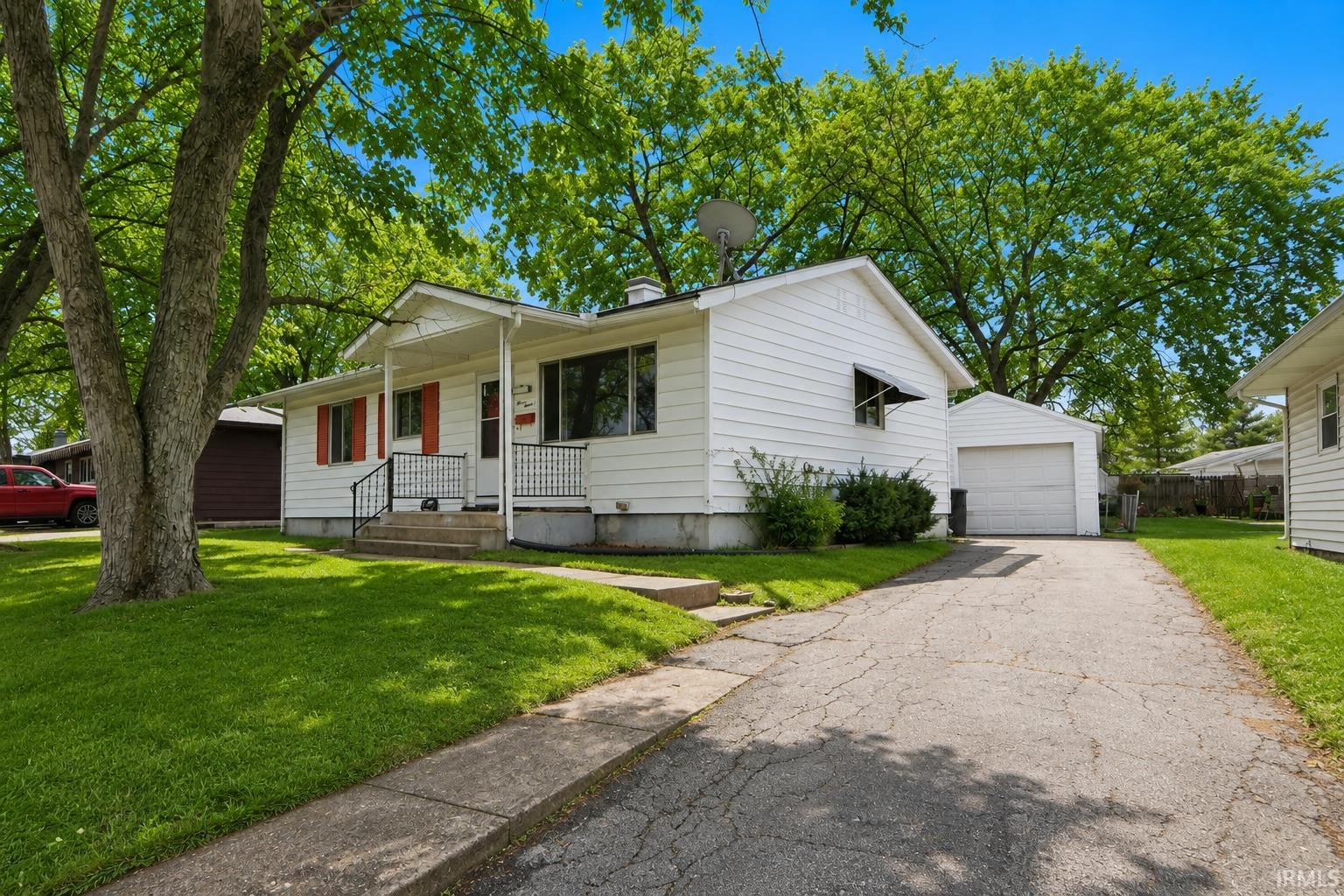 White-sided bungalow with a covered front porch, red window shutters, and a paved driveway leading to a detached garage