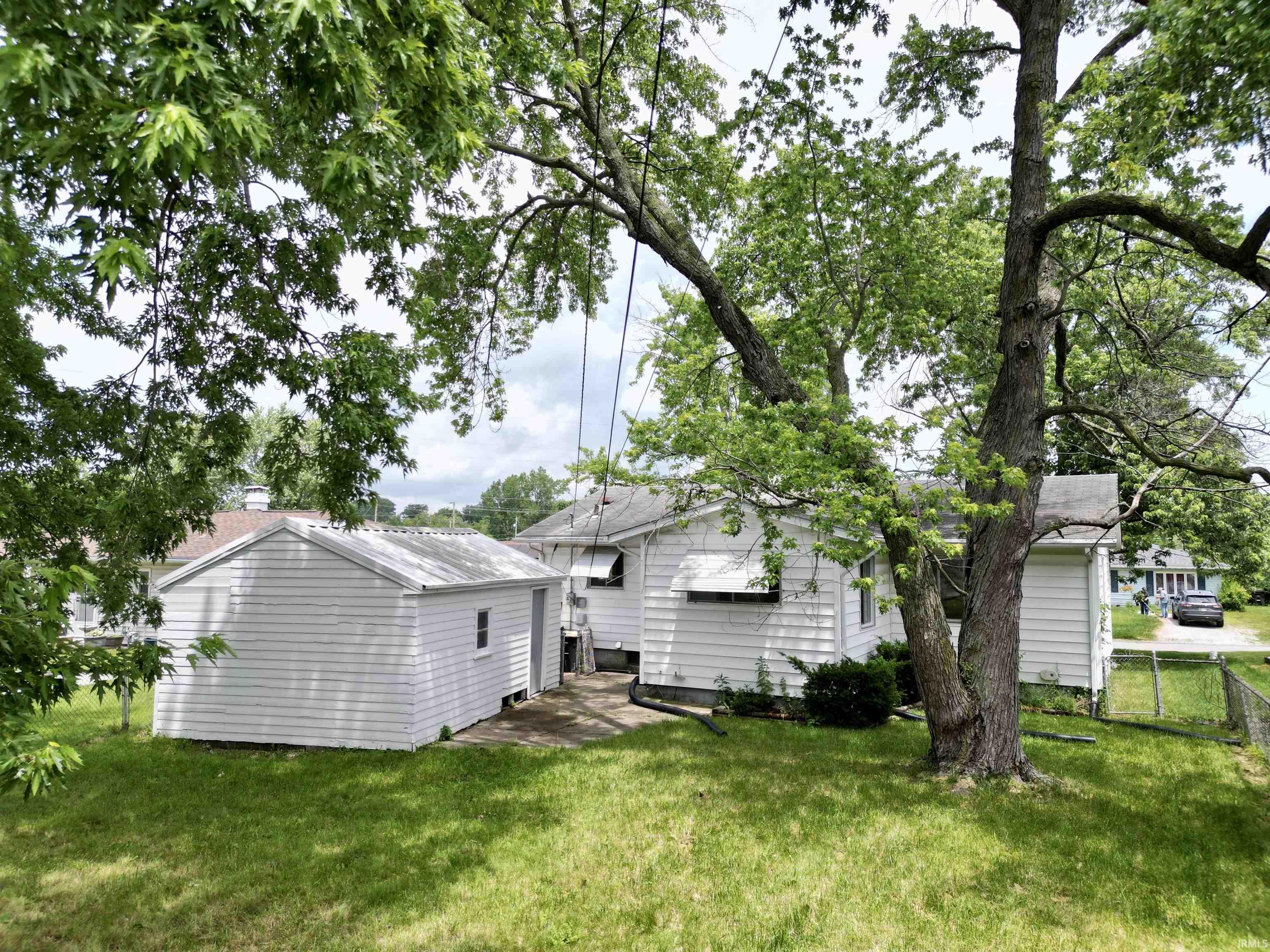 Expansive backyard featuring a detached white storage shed with horizontal siding, a white main residence with horizontal siding, and mature trees providing natural shade