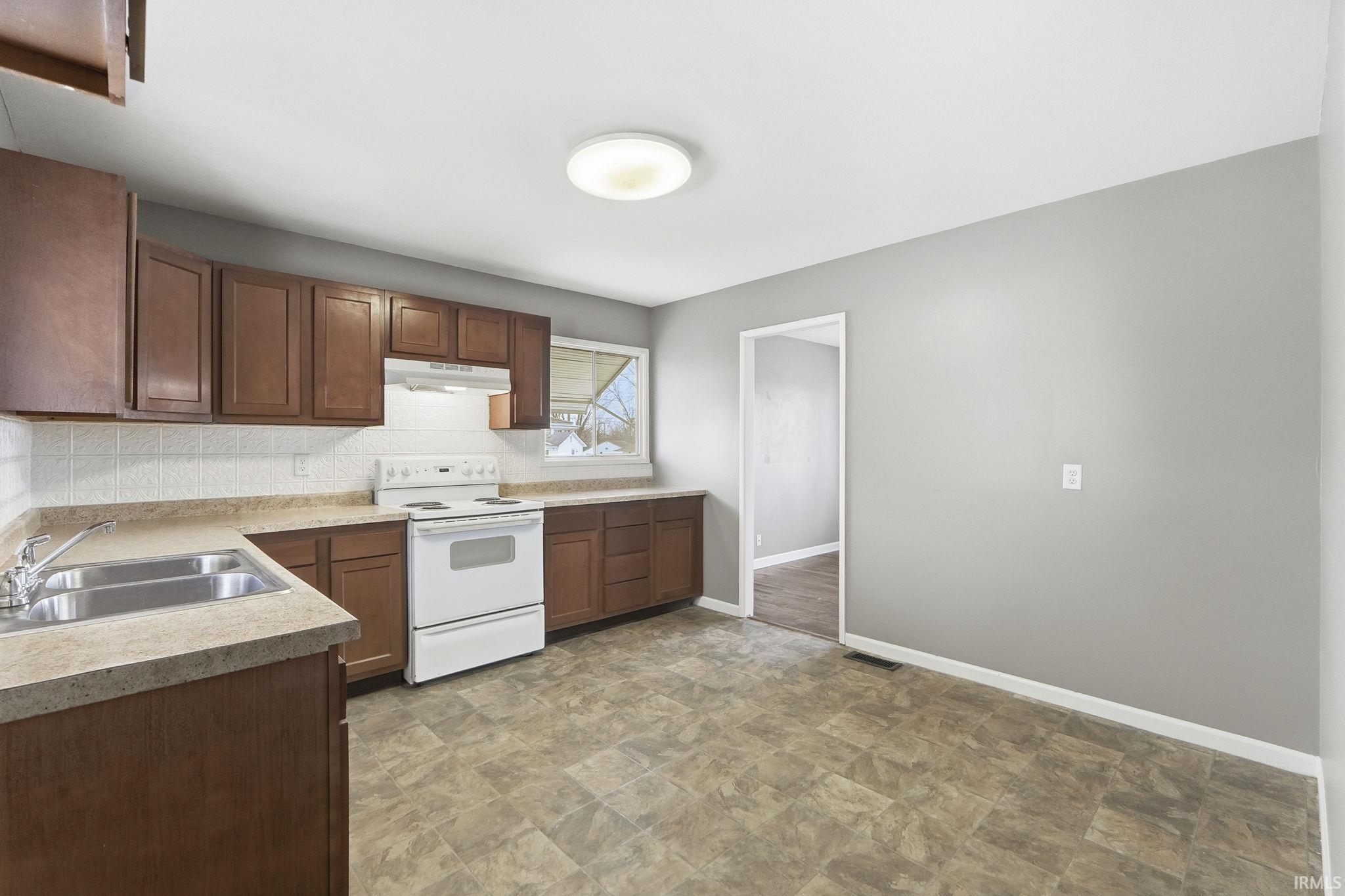 Kitchen featuring electric range, light countertops, stone finish floors, under cabinet range hood, and backsplash