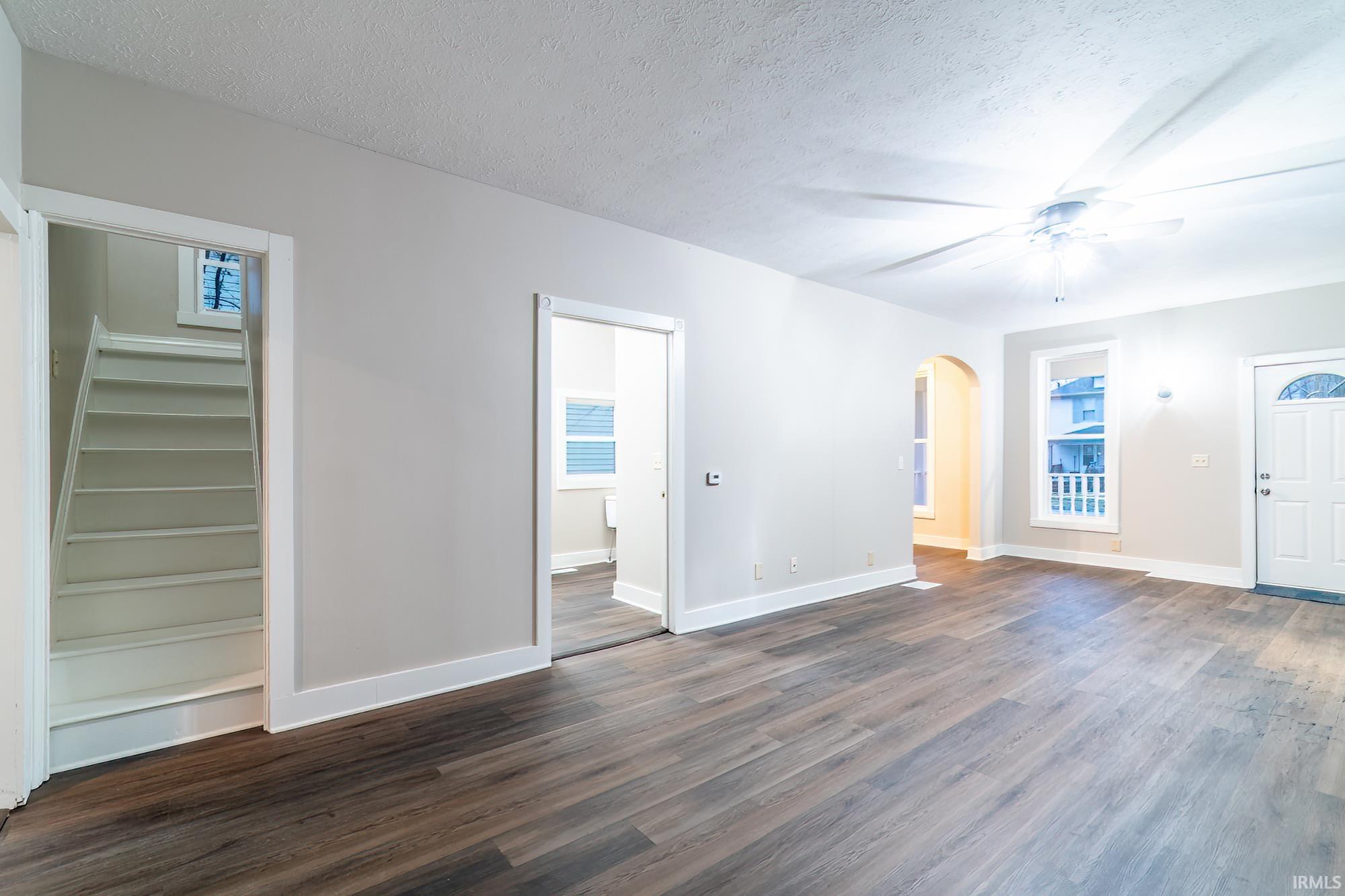 Unfurnished living room with a textured ceiling, dark wood-style floors, arched walkways, ceiling fan, and stairway
