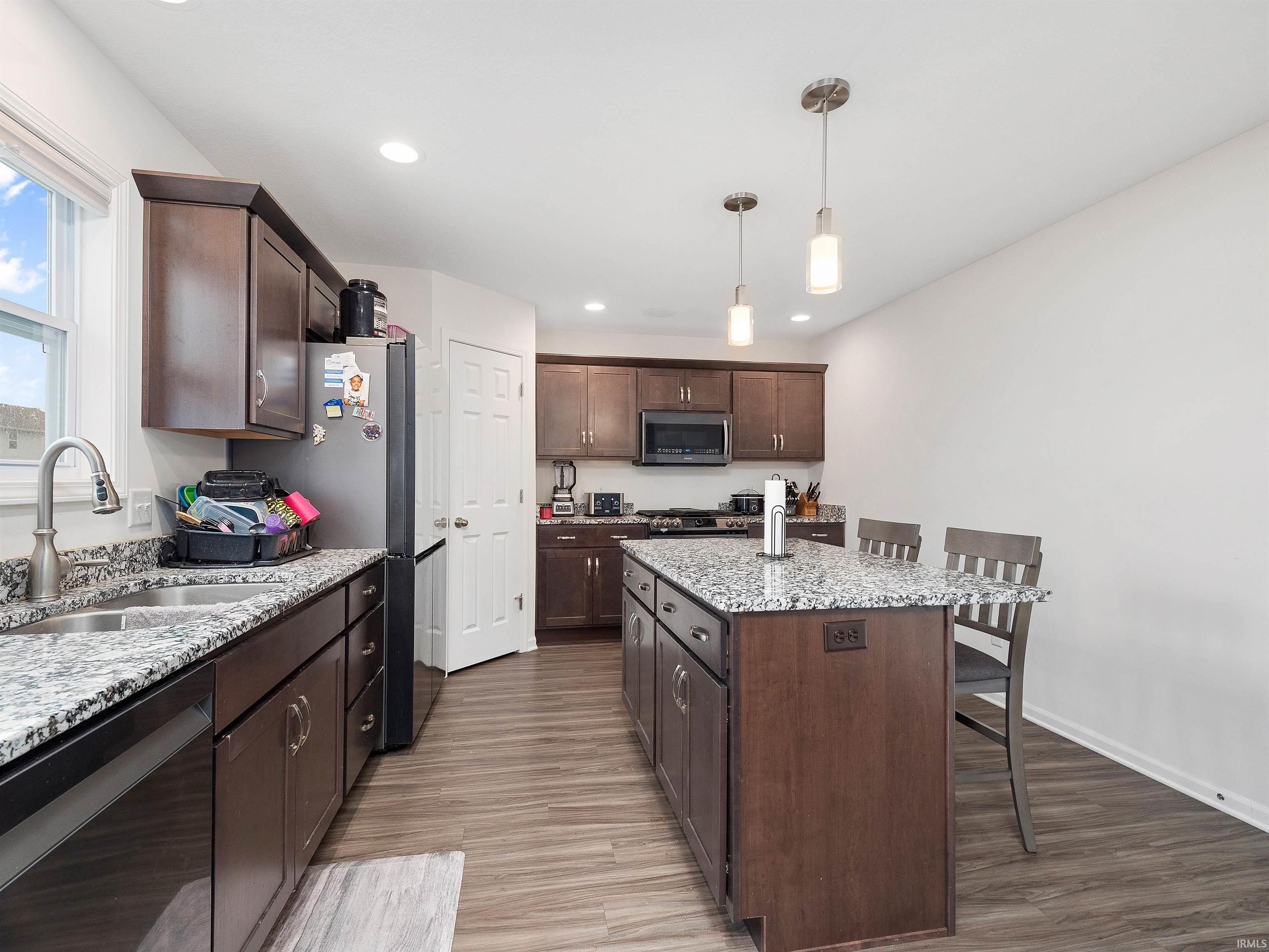 Kitchen featuring dark brown cabinetry, appliances with stainless steel finishes, a kitchen breakfast bar, decorative light fixtures, and light stone counters