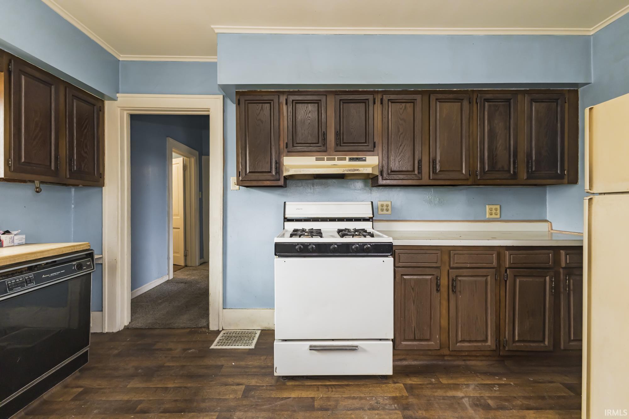 Kitchen featuring white appliances, light countertops, dark brown cabinetry, dark wood-style flooring, and crown molding