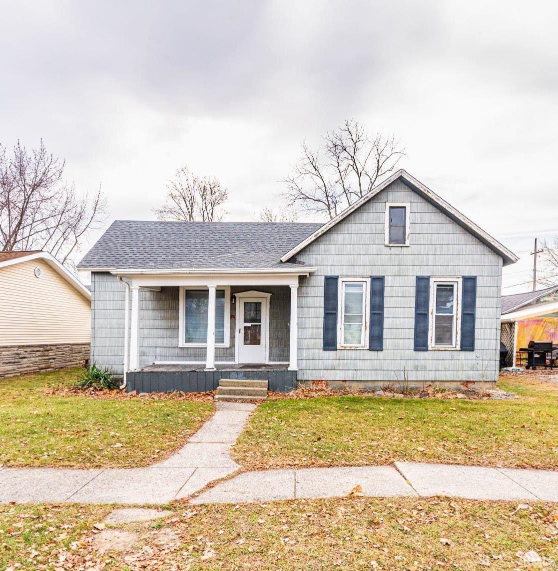 View of front of house with covered porch, a front yard, and roof with shingles