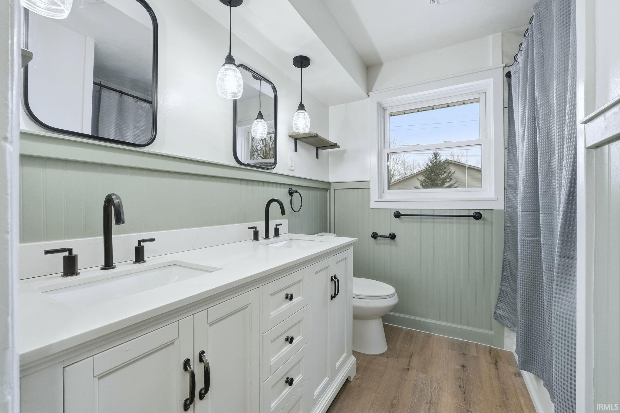 Bathroom with a wainscoted wall, light wood finished floors, a shower with shower curtain, double vanity, and a decorative wall