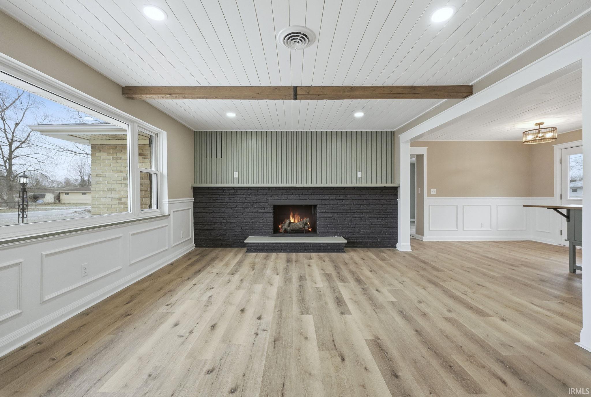 Unfurnished living room featuring a decorative wall, light wood-style floors, a fireplace, a wainscoted wall, and a wooden ceiling with exposed beams