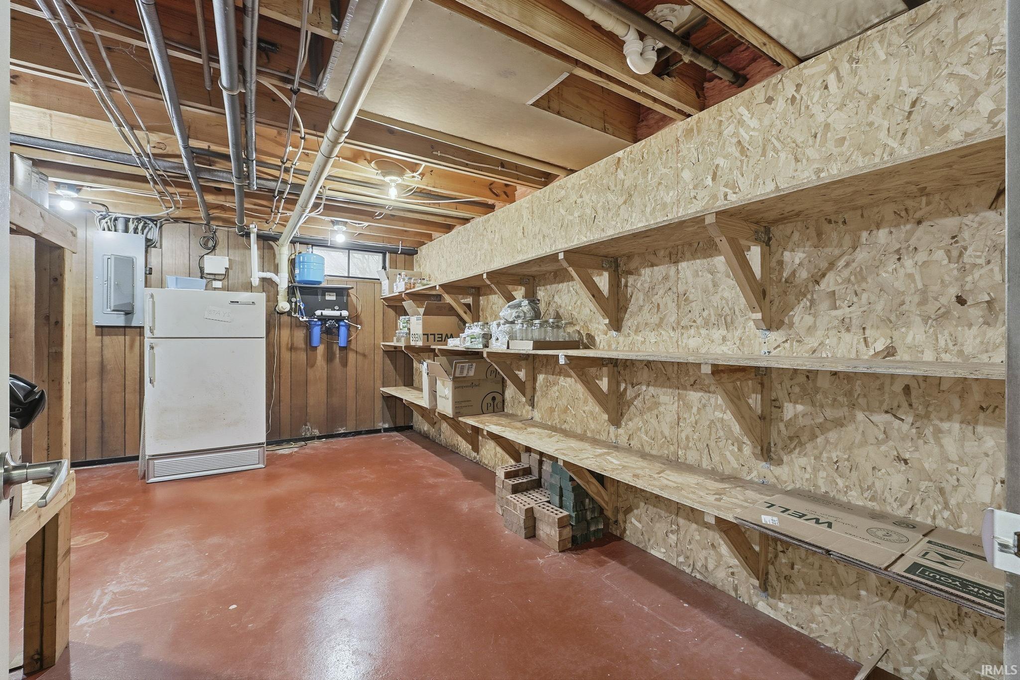 Basement featuring freestanding refrigerator, electric panel, and wood walls