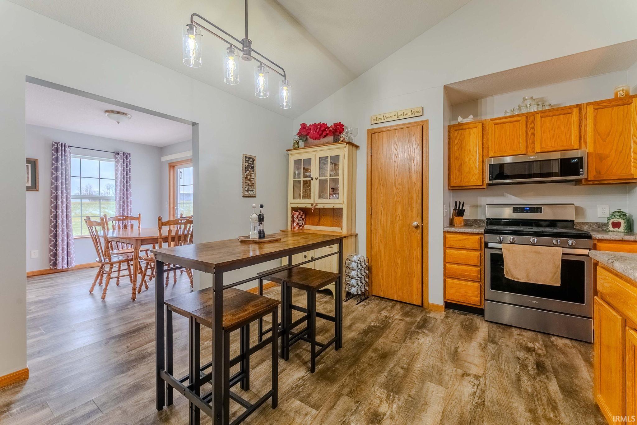Kitchen featuring stainless steel appliances, decorative light fixtures, vaulted ceiling, brown cabinetry, and dark wood-type flooring