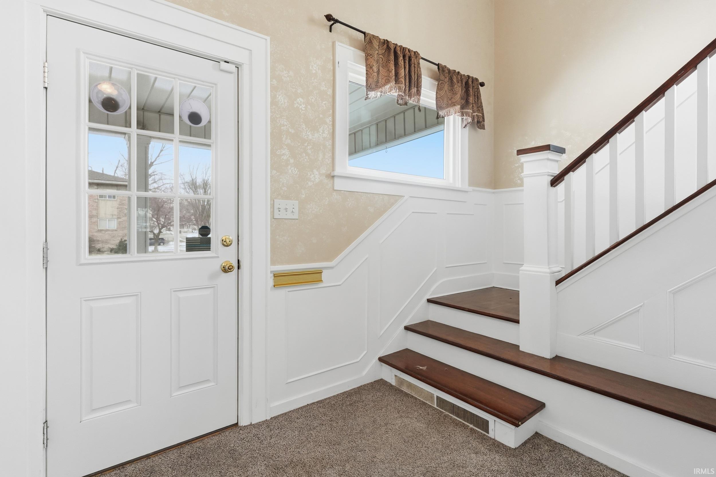 Carpeted entrance foyer featuring a decorative wall, wainscoting, and stairs