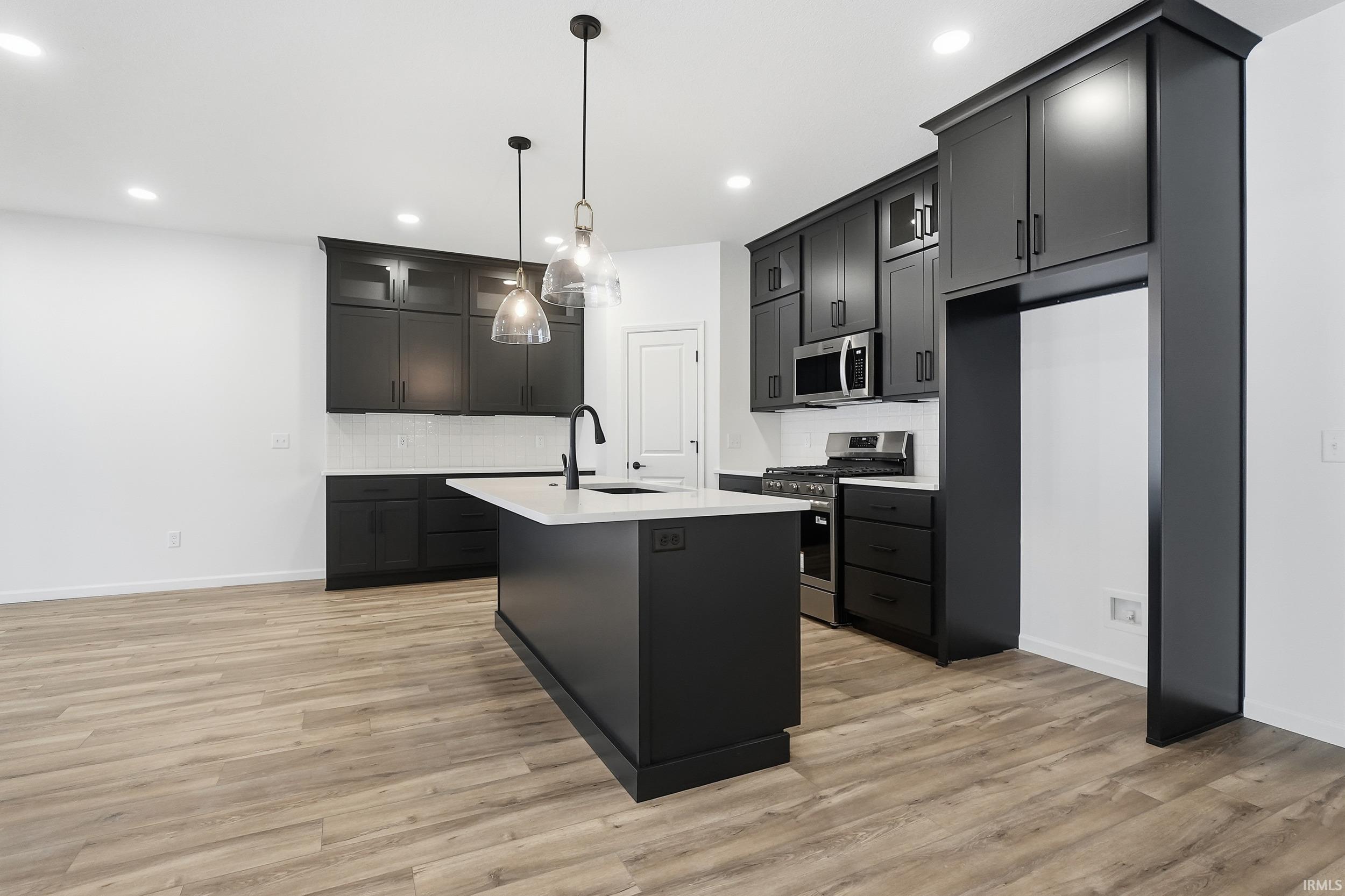 Kitchen featuring stainless steel appliances, backsplash, decorative light fixtures, a kitchen island with sink, and light wood-type flooring