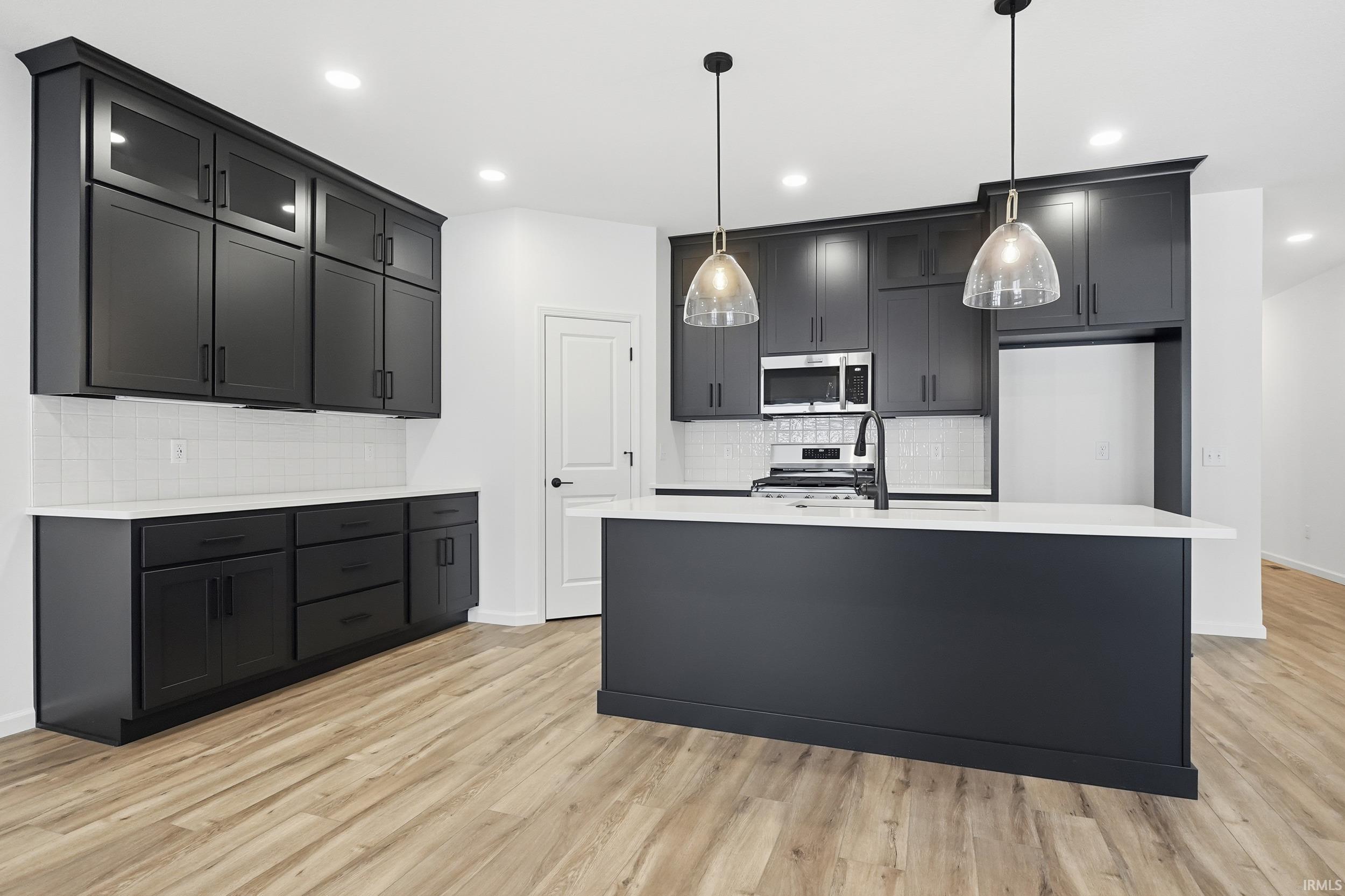 Kitchen featuring decorative light fixtures, appliances with stainless steel finishes, a center island with sink, light wood-style flooring, and light stone counters